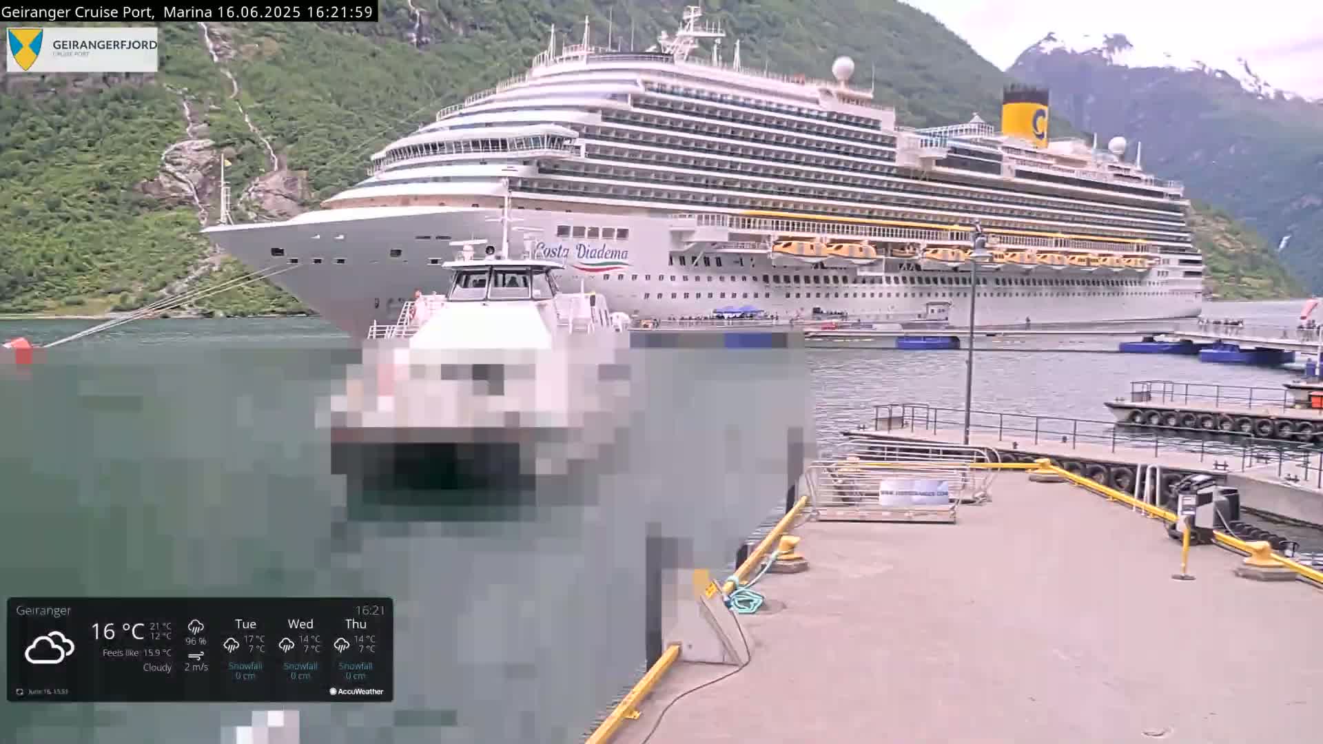 A large cruise ship is docked at a marina, alongside a smaller boat, in a fjord surrounded by mountains under cloudy skies.