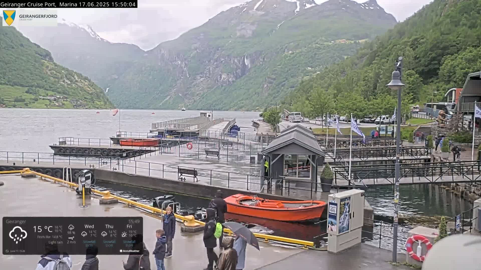 A rainy day at a marina nestled between steep, green mountains, with a small orange boat docked and several people present.