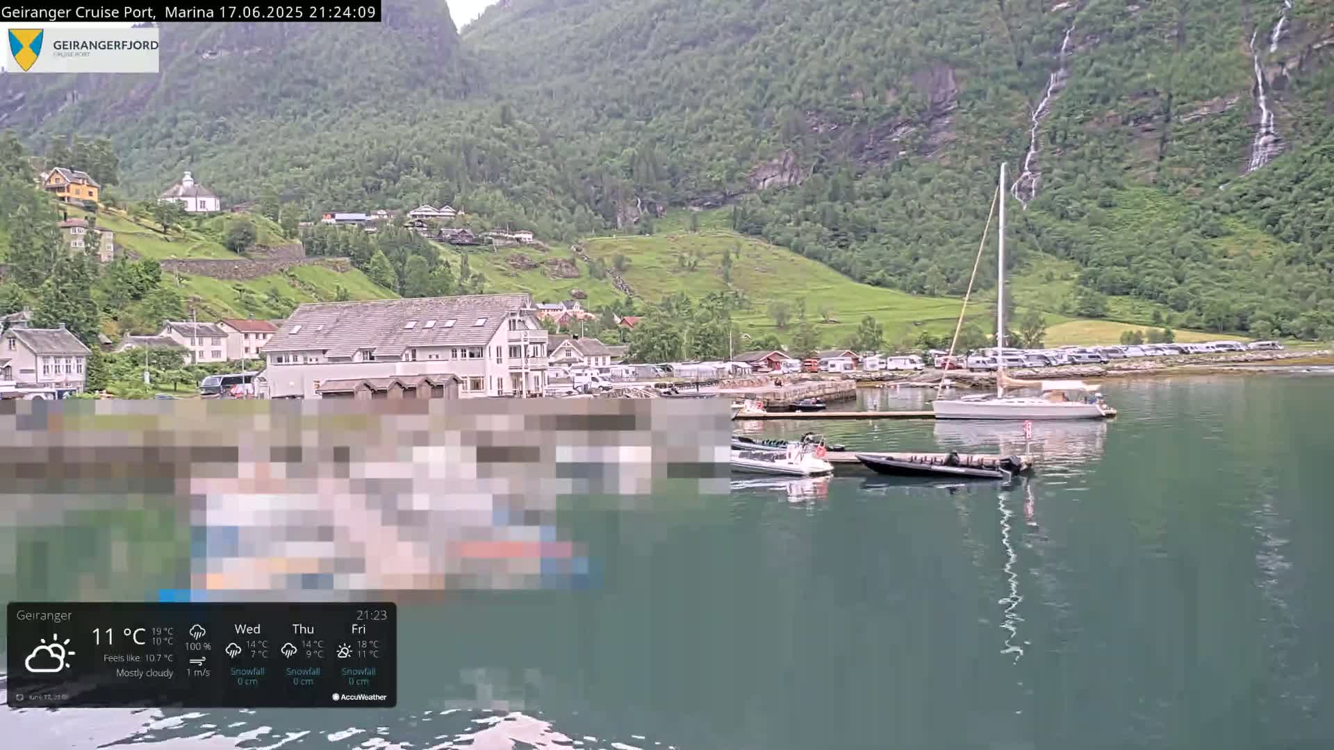 A mostly cloudy waterfront scene shows a marina with several boats, a village nestled against a steep, green hillside with waterfalls cascading down the mountains.