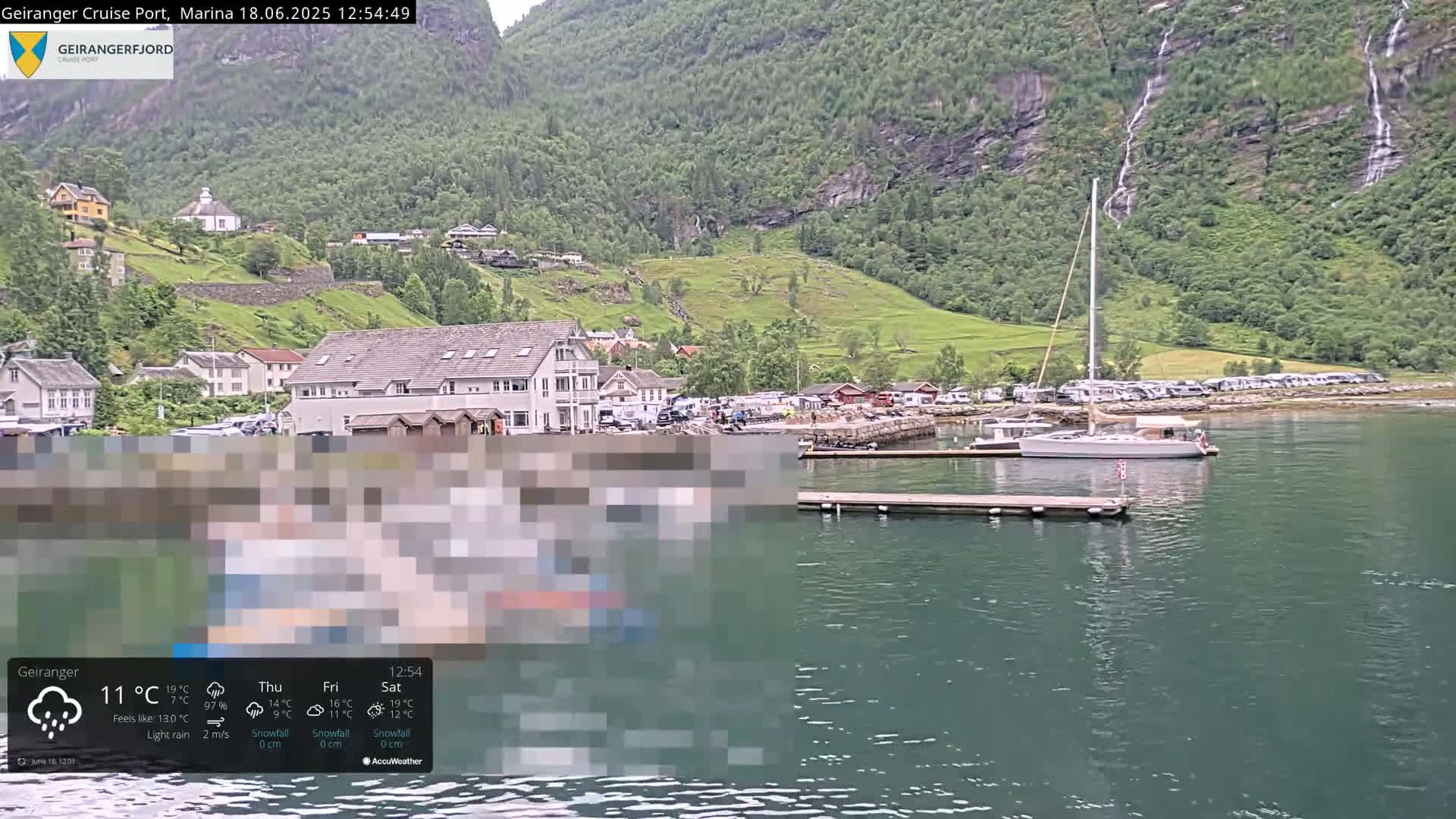 A sailboat is moored in a calm, green fjord near a village nestled against a verdant hillside with waterfalls cascading down its rocky face under light rain.