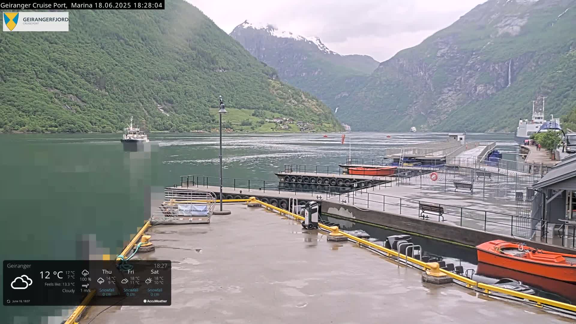 A wet dockside area with several boats, a ferry in the distance, and a fjord surrounded by steep green mountains under a cloudy sky.