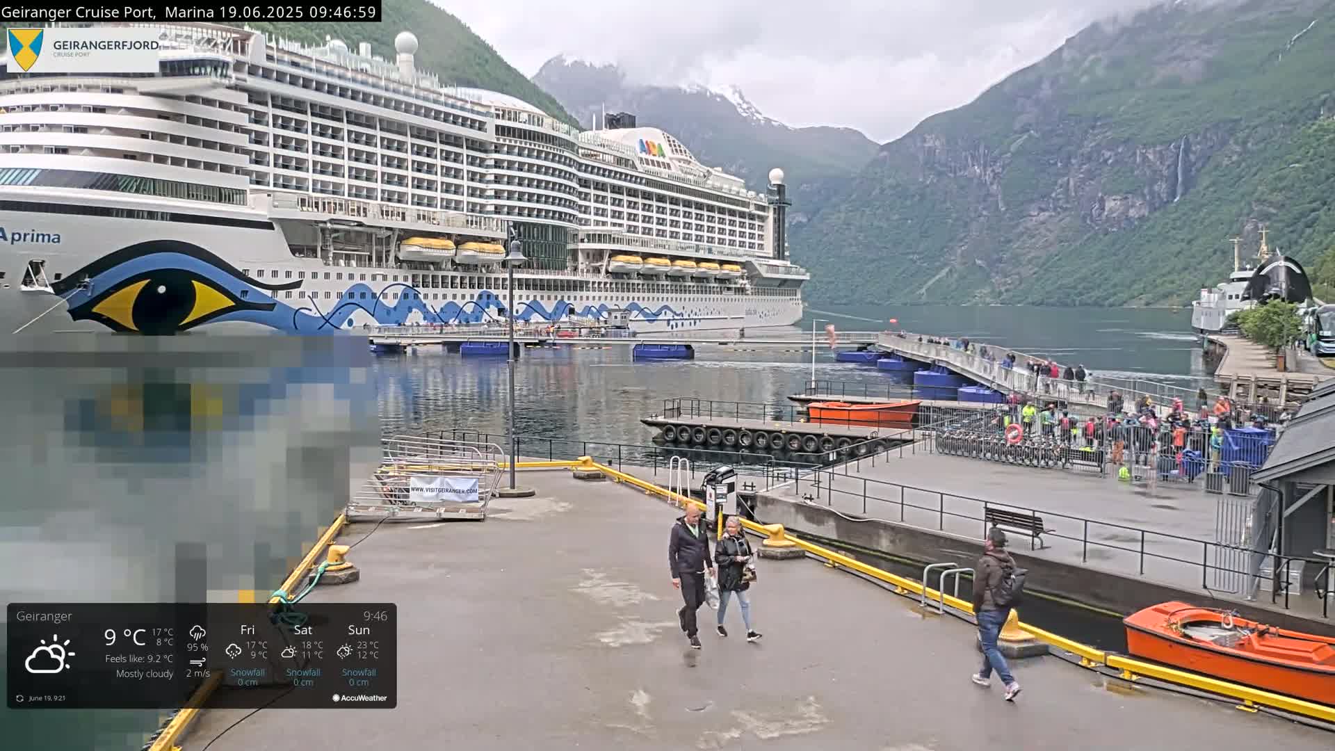A large cruise ship is docked at a port in a fjord, under mostly cloudy skies, with people walking on the pier and smaller boats nearby.