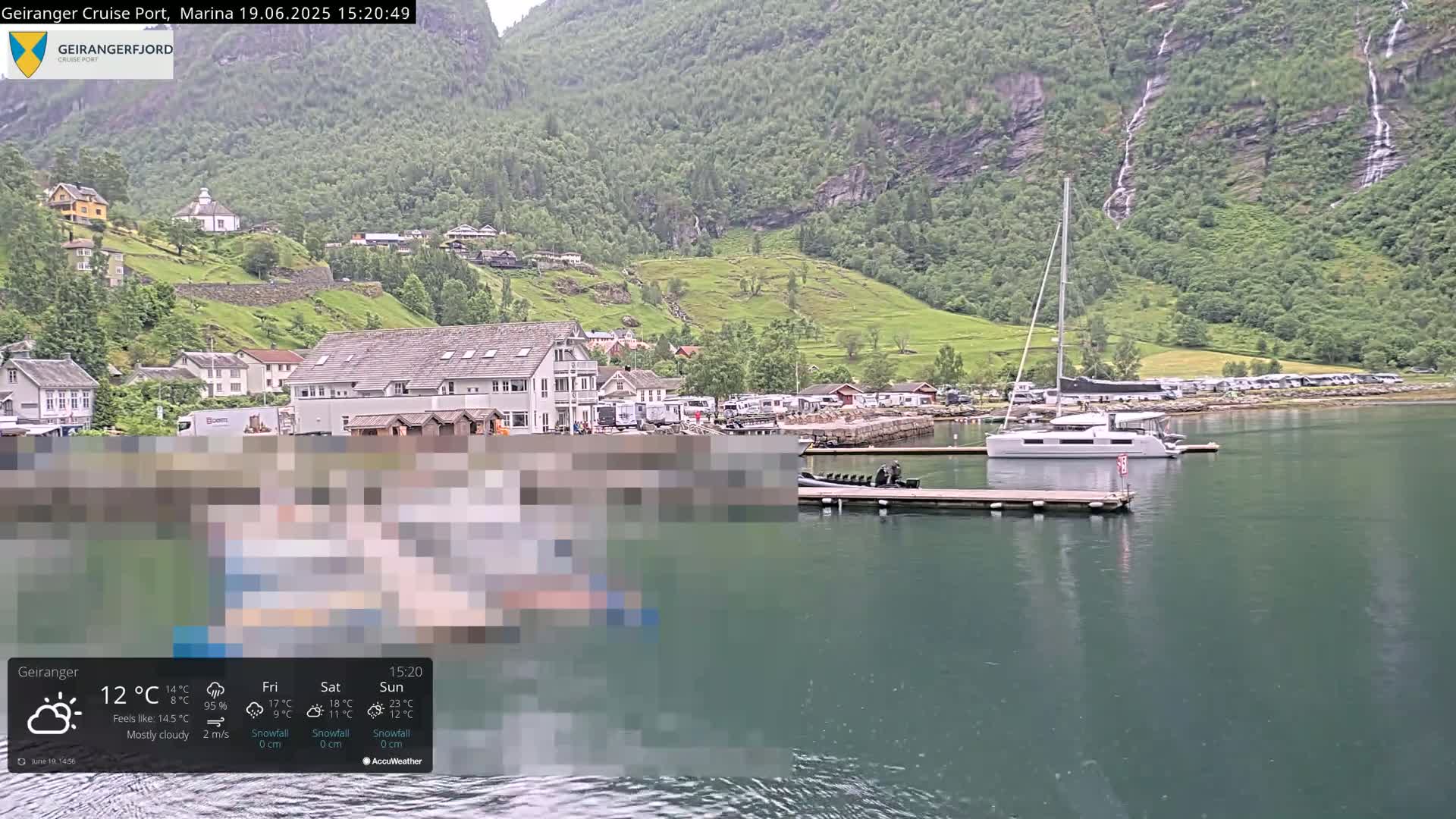 A marina with boats and buildings nestled beside a calm body of water, backed by lush green hills and waterfalls under mostly cloudy skies.