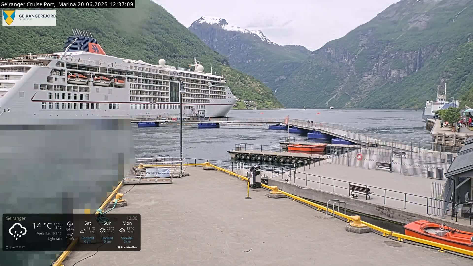 A large cruise ship is docked at a marina in a fjord, with light rain falling and mountains in the background.