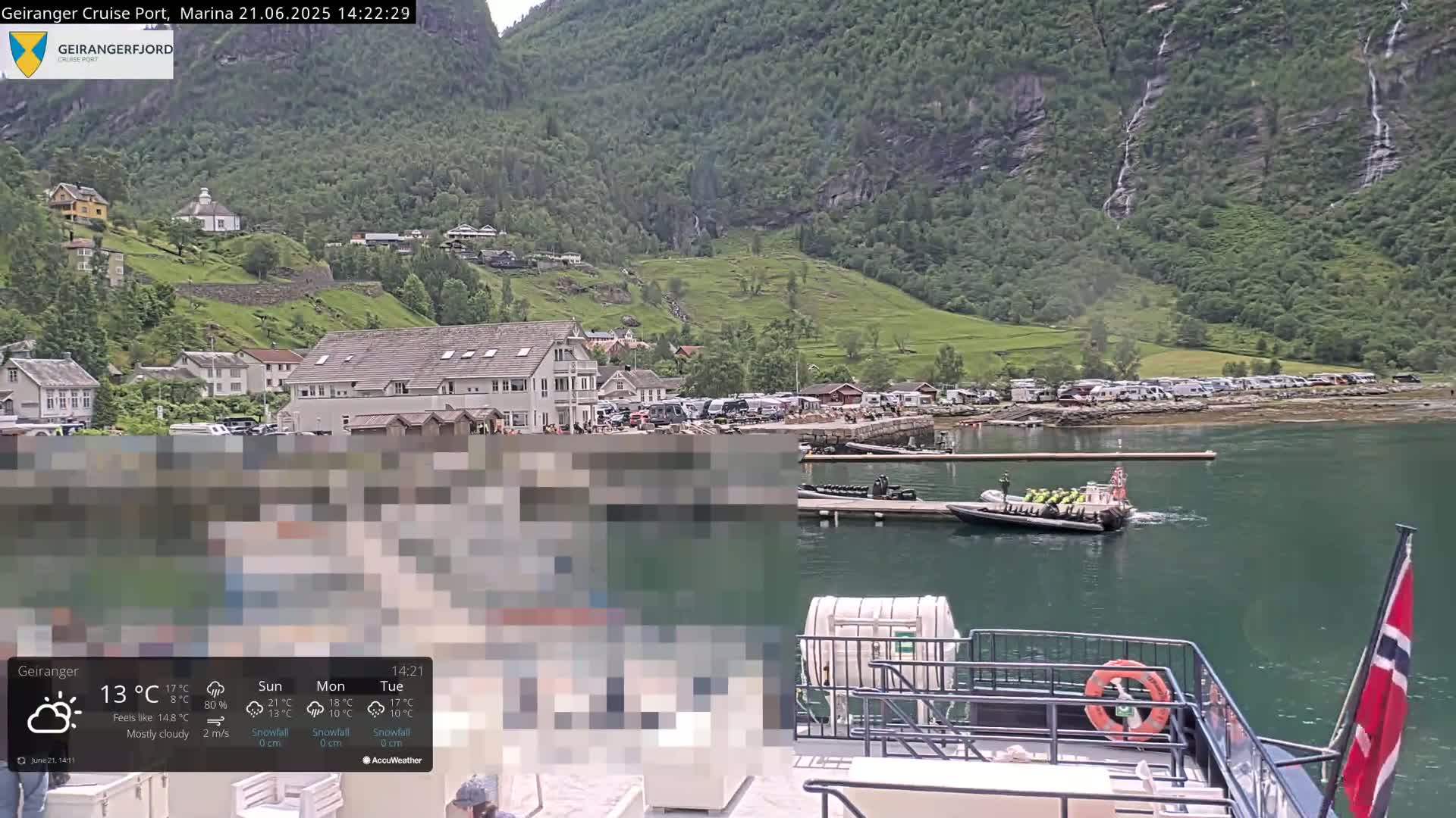 A mostly cloudy day shows a harbor village nestled against a steep, green mountainside with several waterfalls cascading down, as seen from the back of a boat.