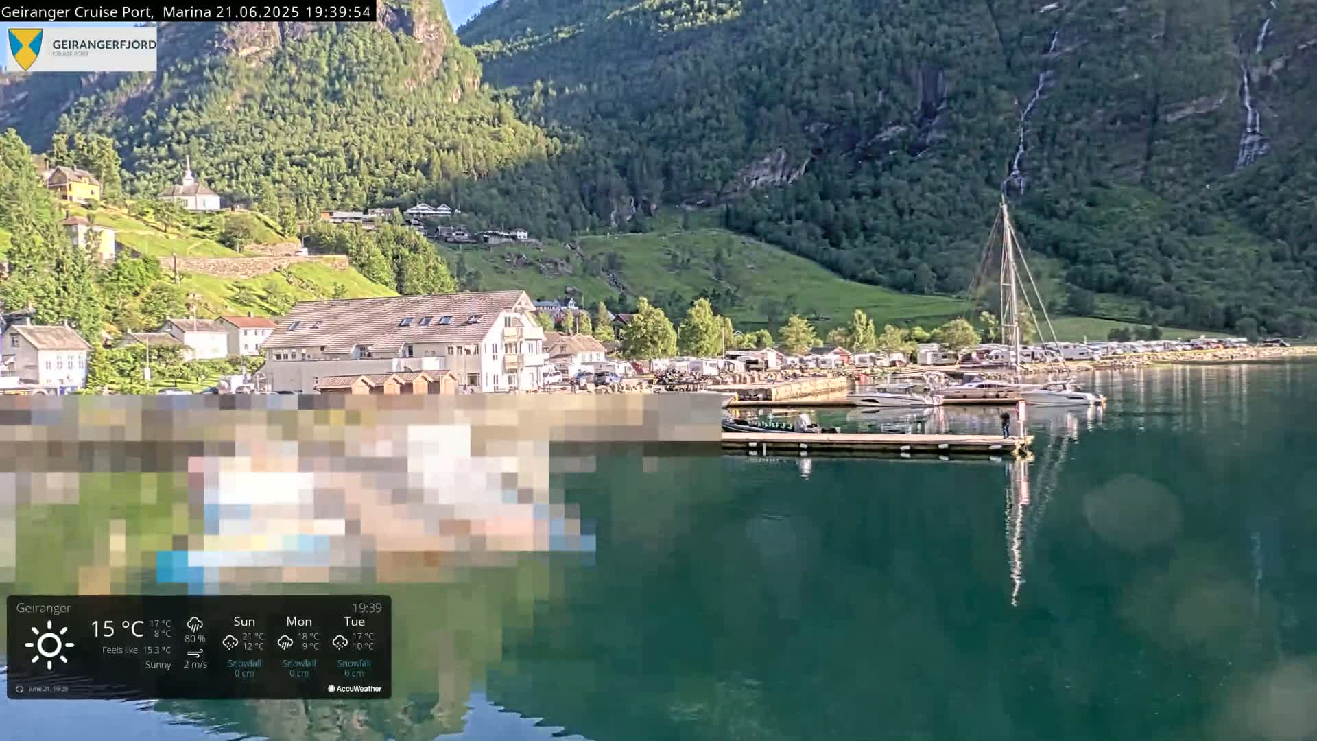 A sunny harbor scene shows a calm body of water with boats docked near buildings and a hill covered in lush green vegetation.