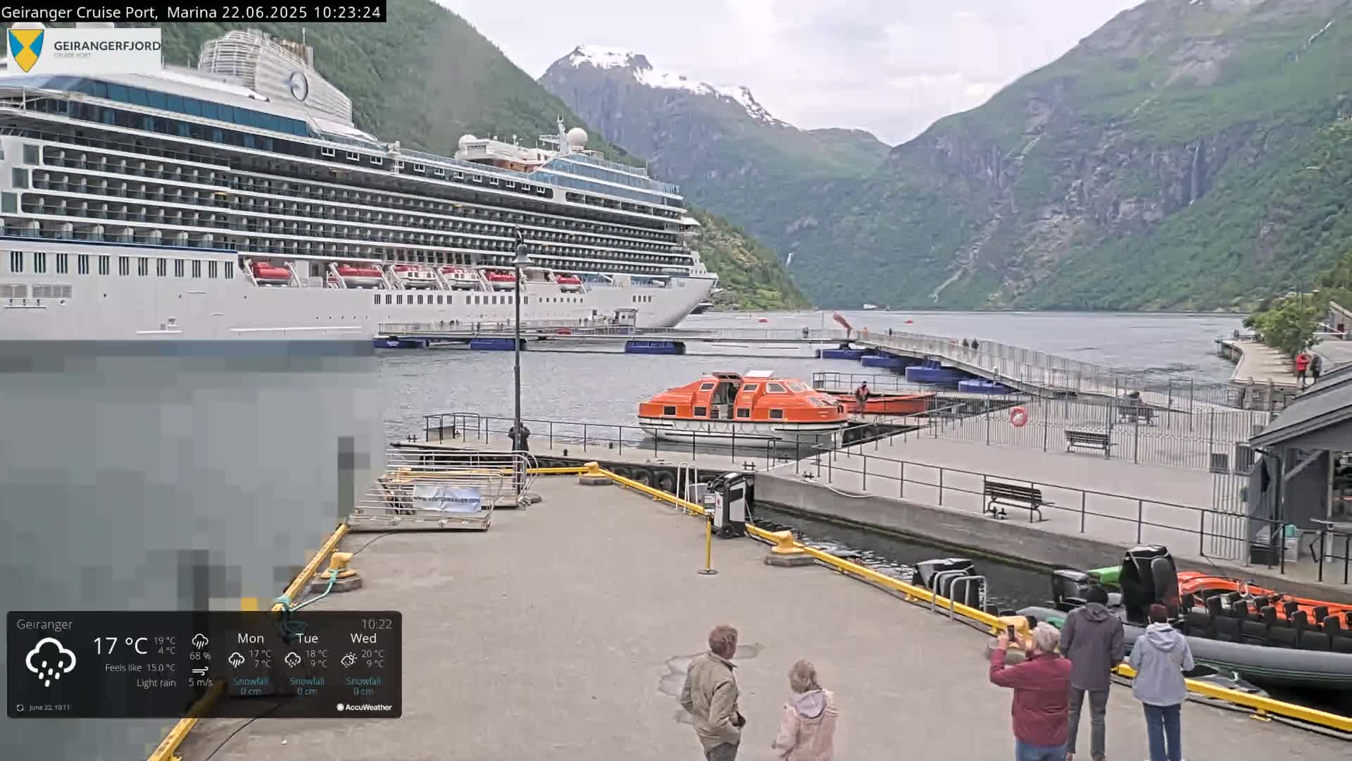 A large cruise ship is docked at a marina, with a smaller orange lifeboat nearby, under overcast skies and light rain in a fjord surrounded by mountains.