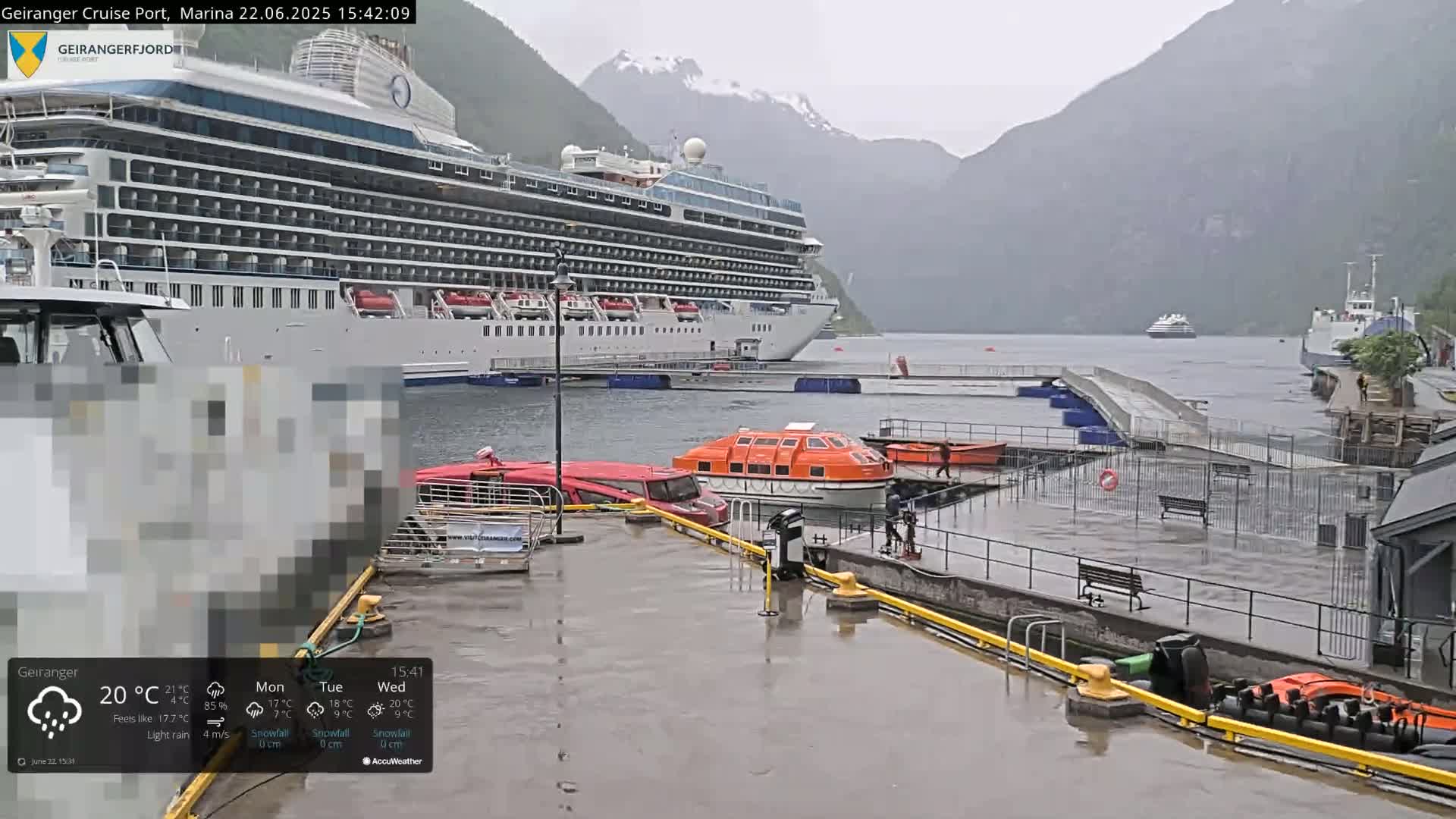 A large cruise ship is docked at a port in a fjord, surrounded by rain-soaked docks and mountains under a light rain.