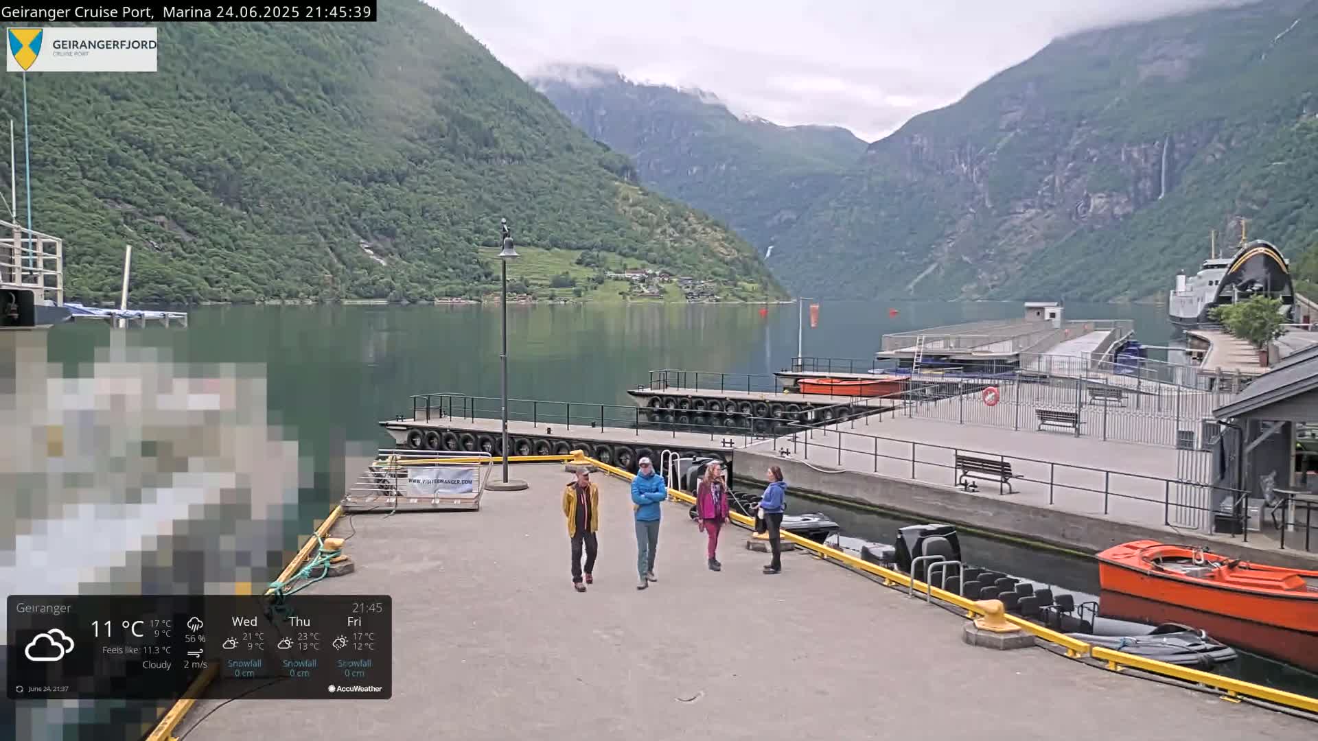 A cloudy day at a marina nestled between steep, green mountains, with several small boats and a larger vessel moored nearby, and four people walking on the dock.