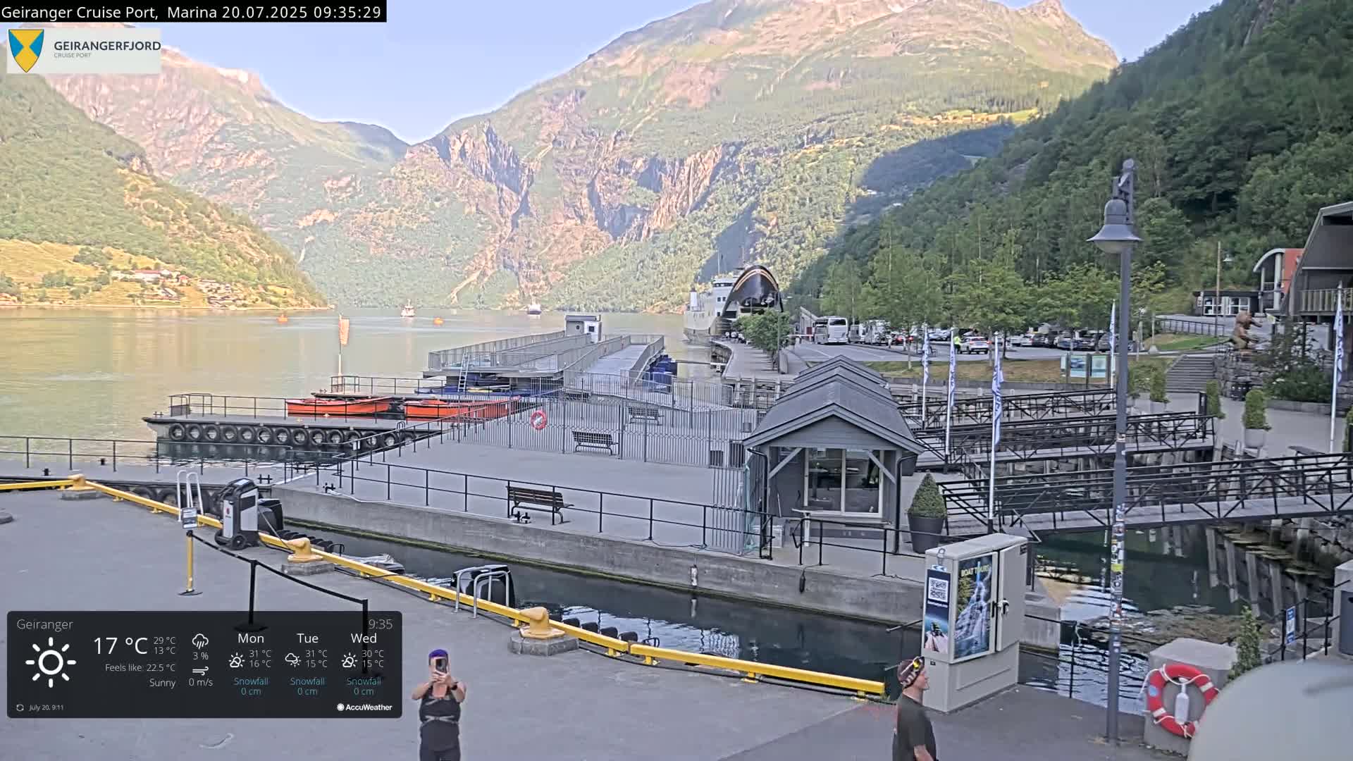 A sunny view of a marina nestled between steep, green mountains, with several boats docked and a ferry visible in the distance.