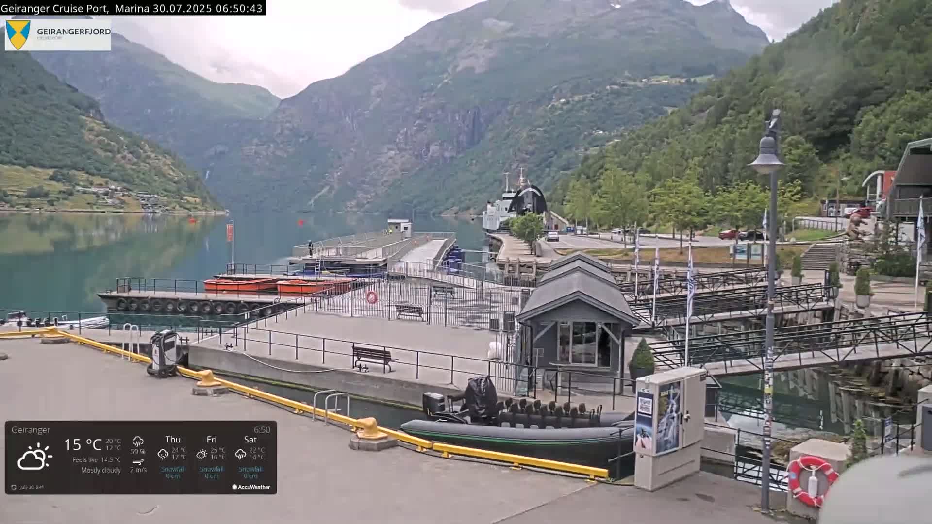 A sunny view of a marina nestled between steep, green mountains, with several boats docked and a ferry visible in the distance.