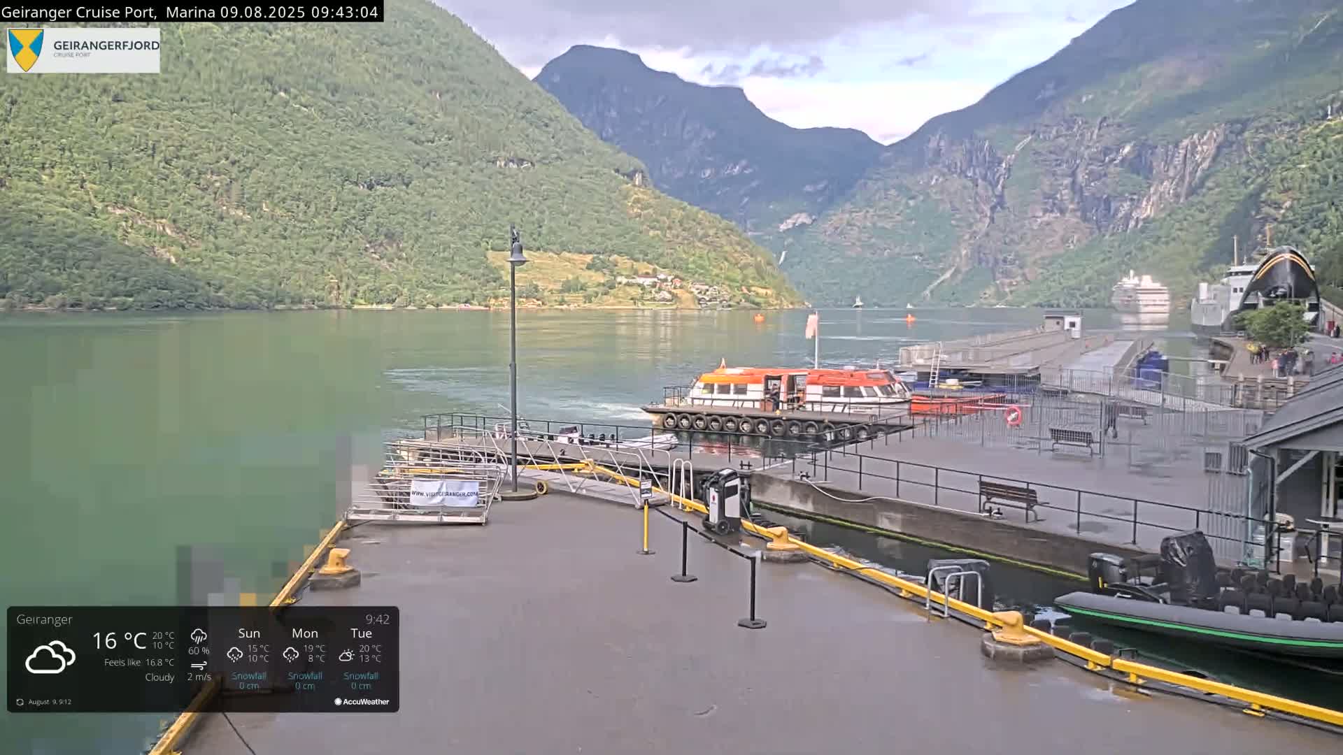 A cloudy day at a fjord marina shows several boats docked, including a large orange passenger boat, with steep green mountains in the background.