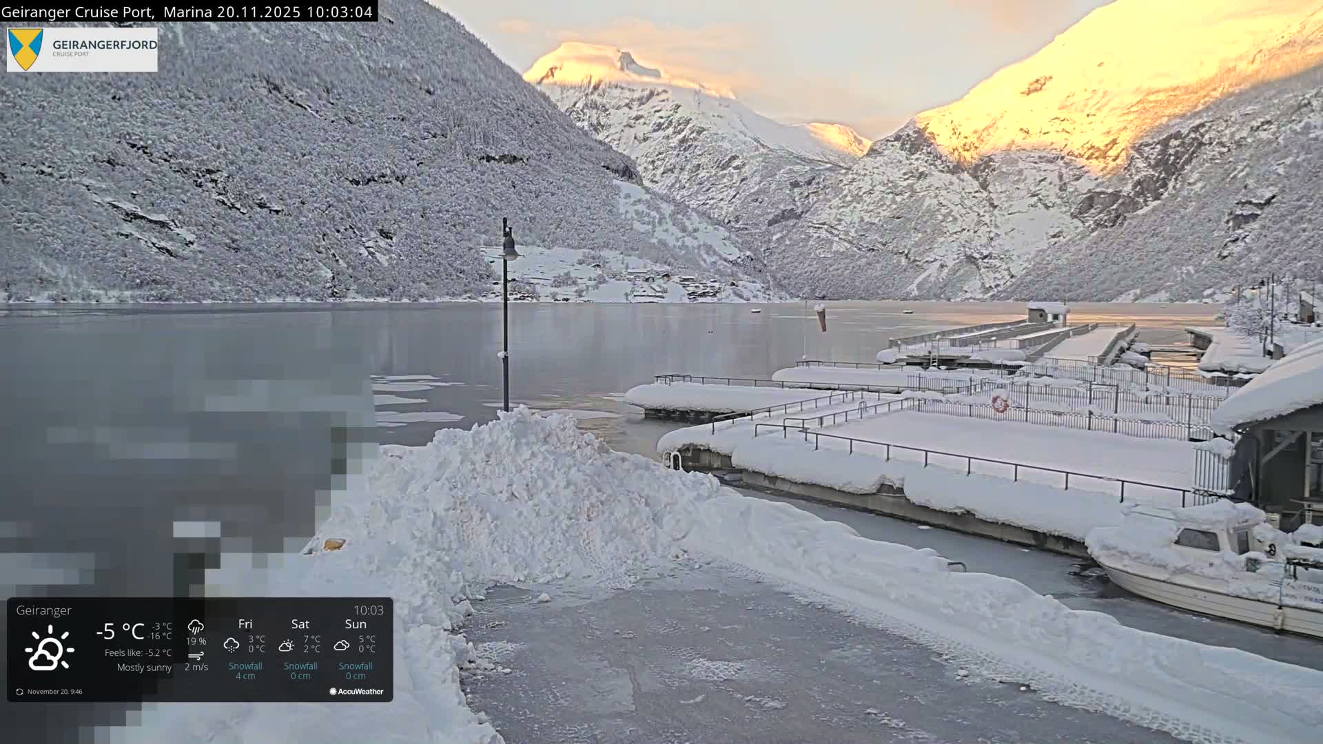 A tranquil winter scene captures a snow-covered marina with docked boats on partially icy water, nestled among towering, snow-dusted mountains, under bright, cold conditions with sunlight illuminating the peaks.