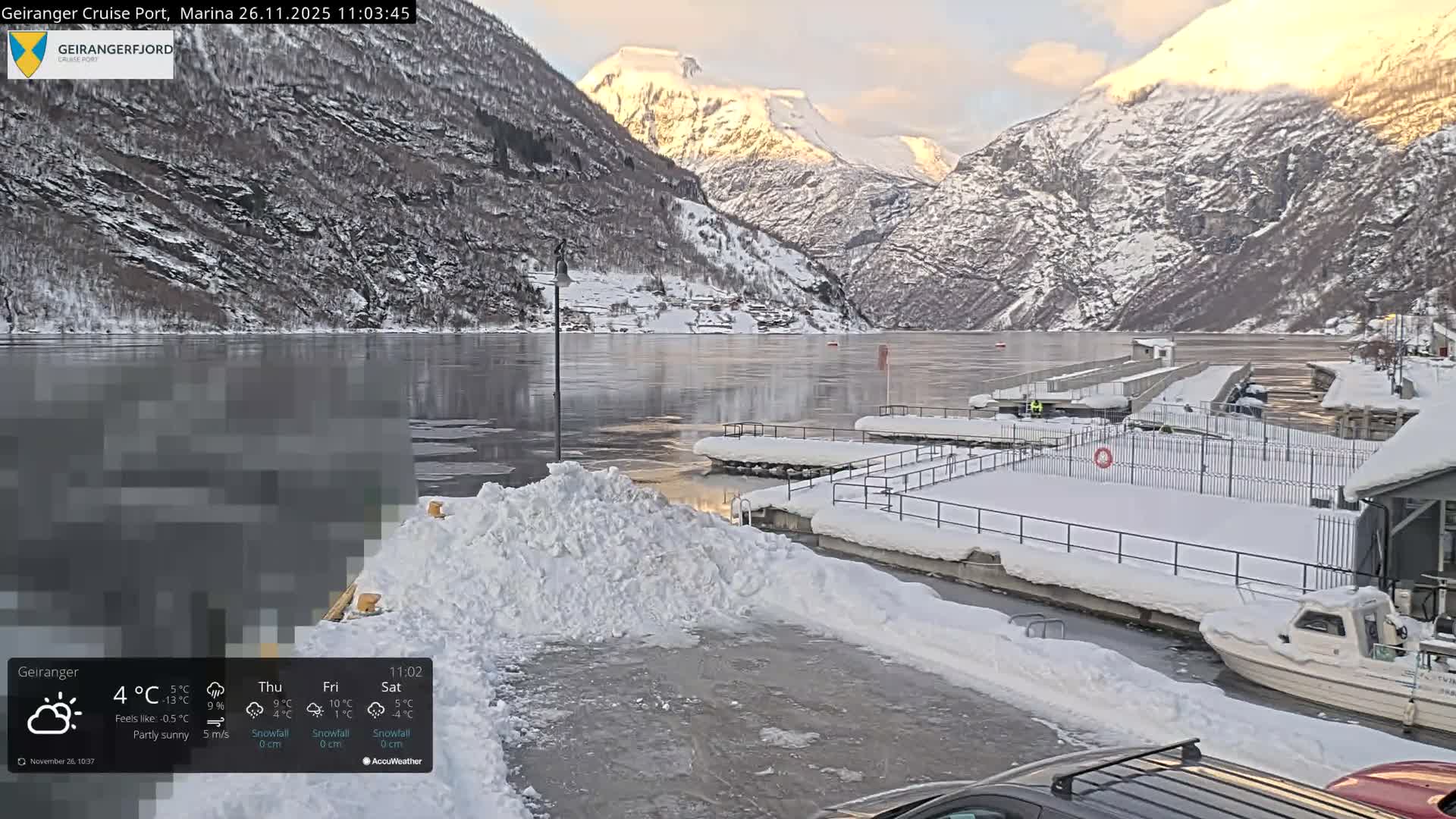 A serene winter scene depicts a snow-covered fjord with partially frozen water reflecting the sky, flanked by majestic snow-capped mountains illuminated by the sun, with snow-laden docks and a small boat in the foreground under partly sunny skies.