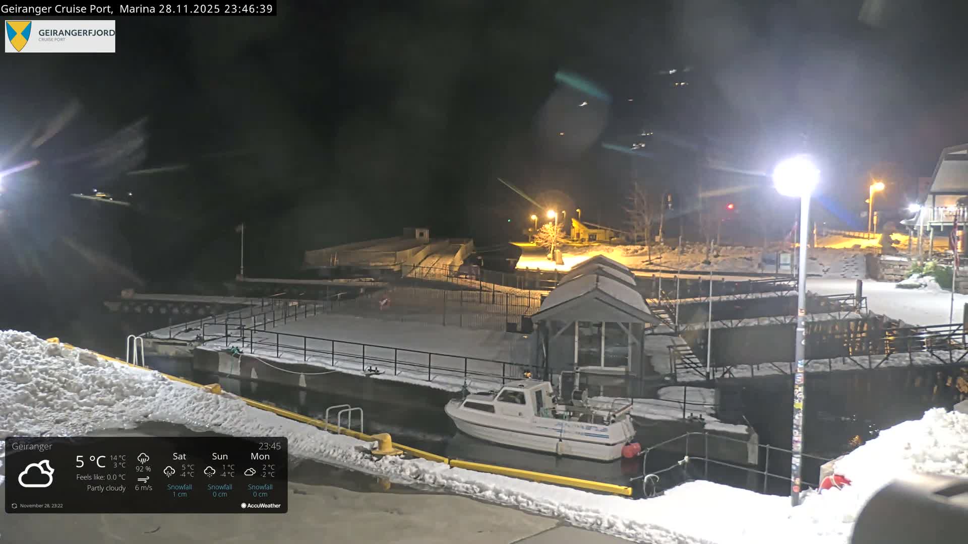 A tranquil, snow-covered harbor at night reveals a white boat docked in dark water beside snowy piers and structures, illuminated by streetlights under clear, cold winter conditions.