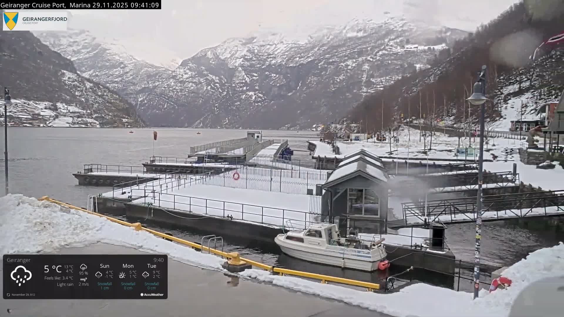 A snow-covered marina with docks and a small white boat is nestled amidst towering, snow-dusted mountains under a bright, overcast sky.