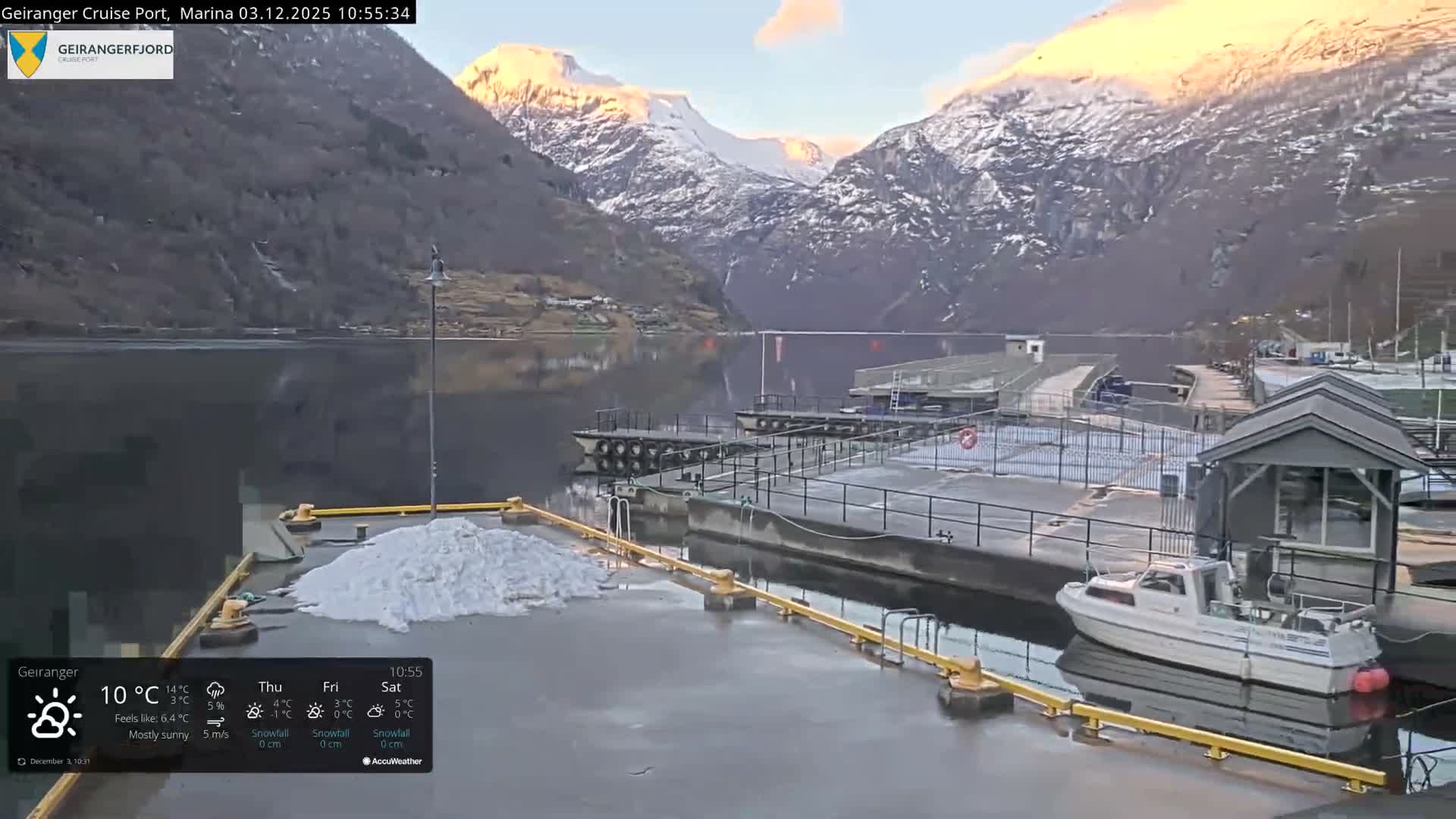 A tranquil winter scene shows a calm marina with docks, a small boat, and scattered snow on the ground, all set against a backdrop of majestic snow-capped mountains under a clear sky, with soft light illuminating some distant peaks.