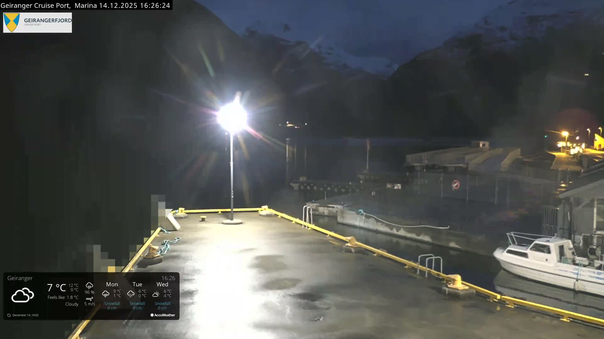 A tranquil winter scene shows a calm marina with docks, a small boat, and scattered snow on the ground, all set against a backdrop of majestic snow-capped mountains under a clear sky, with soft light illuminating some distant peaks.
