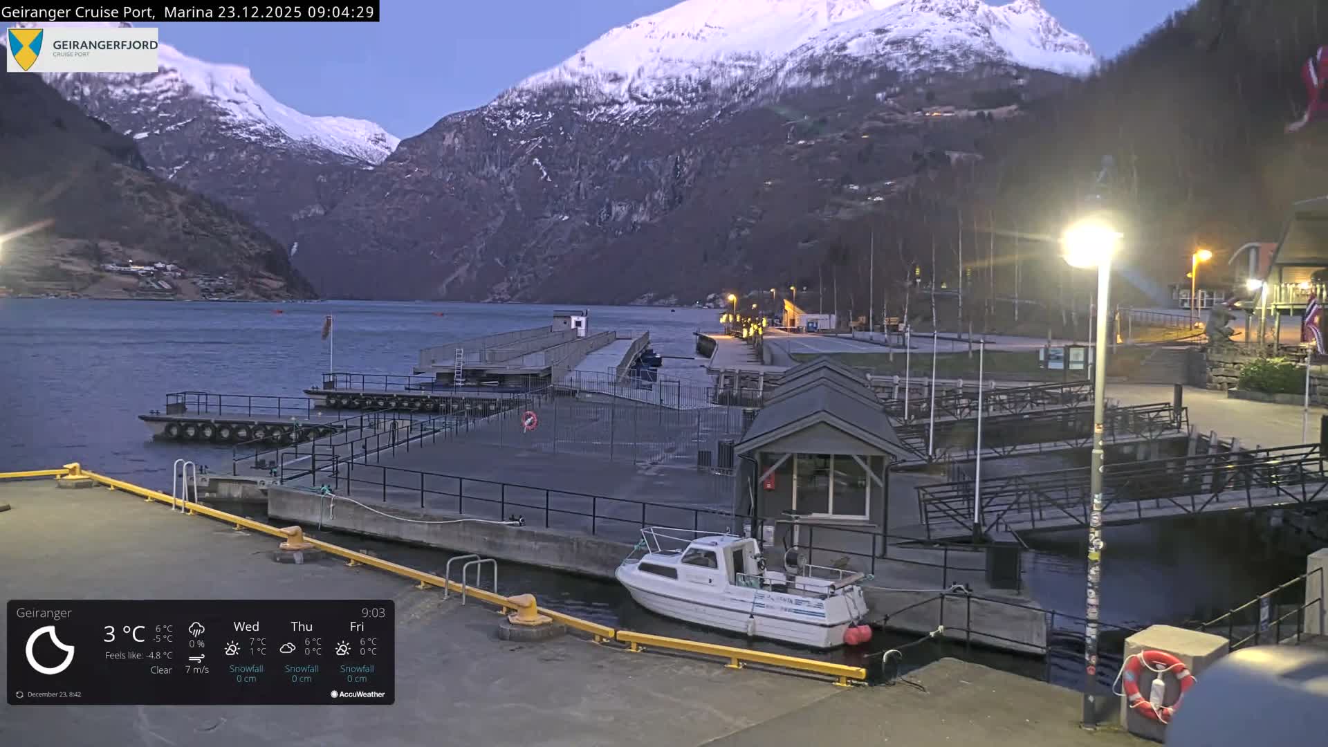 A serene, cold twilight scene shows snow-capped mountains towering over a calm fjord with several docks, a white boat, and illuminated buildings along the water's edge.