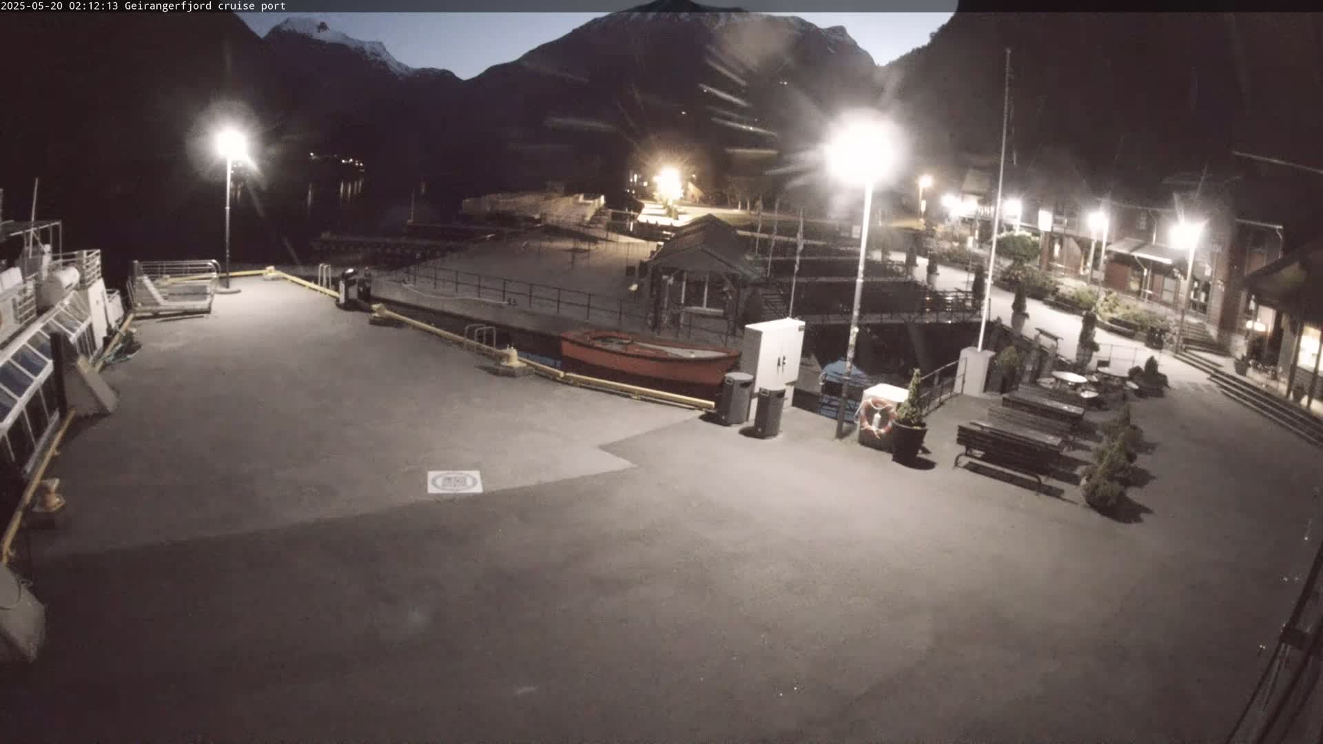 A nighttime view of a cruise port shows a mostly empty plaza with a docked ship, small boats, and benches, all illuminated under artificial light against a backdrop of dark mountains.