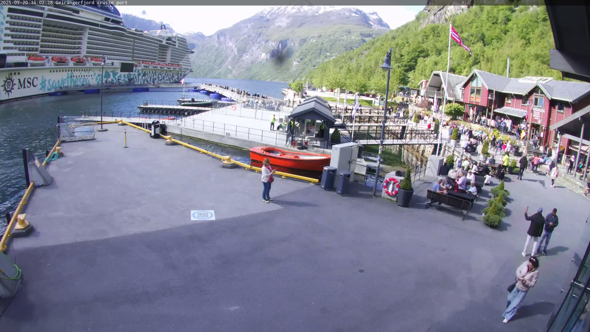 A large cruise ship is docked at a port, with many people gathered on the dock and surrounding buildings on a sunny day amidst a mountainous landscape.
