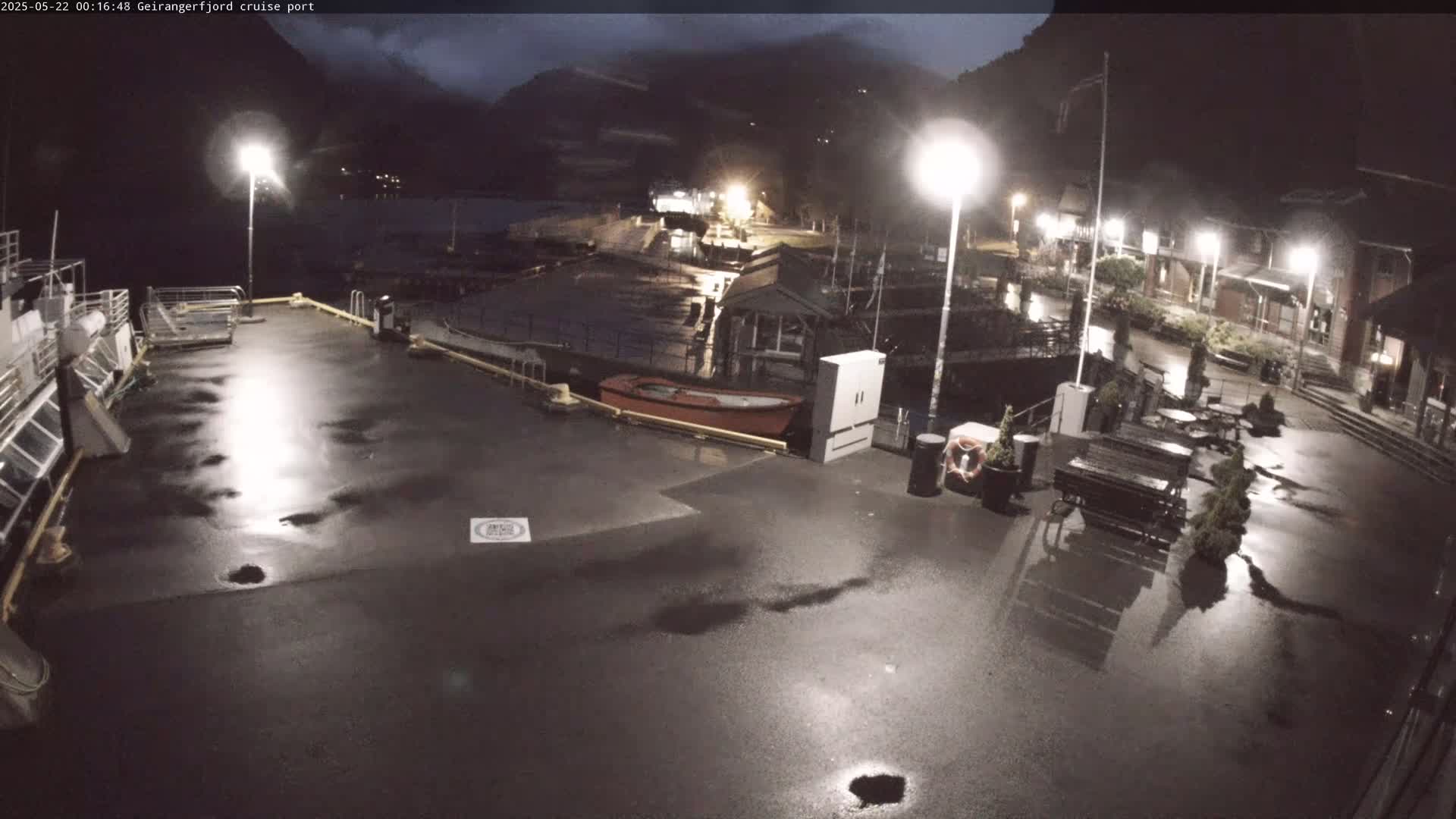 A wet, nighttime cruise port shows a mostly empty dock with a few boats and buildings under artificial light, with dark mountains in the background.