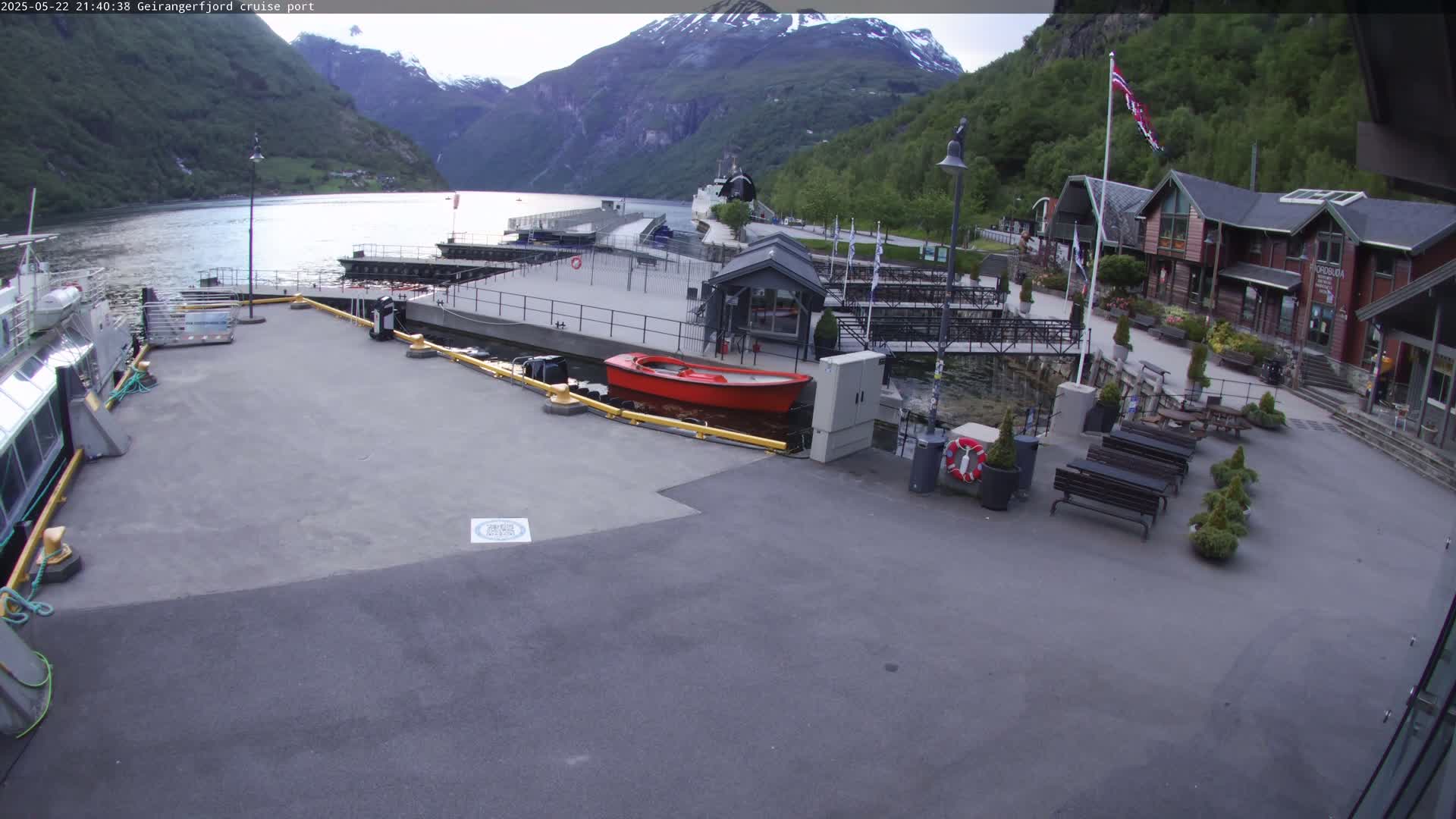 A cruise ship port with a red boat in the water, several buildings, and some seating areas, all situated against a backdrop of mountains under an overcast sky.