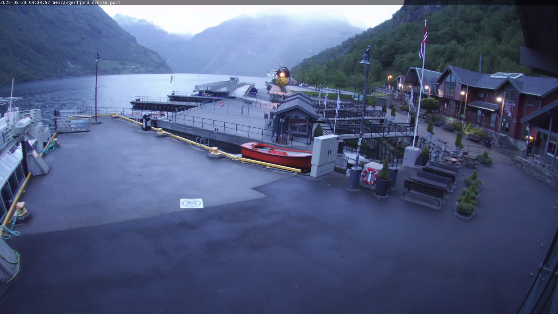 A cruise ship port with calm water, mountains in the background, a small red boat, and several buildings under an overcast sky.