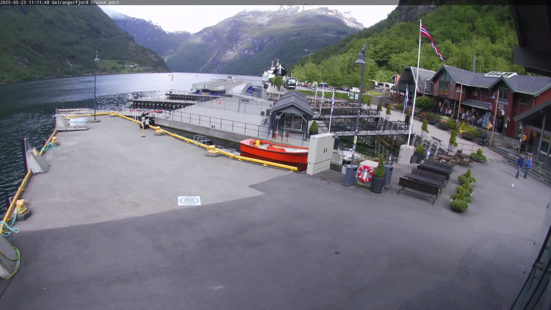 A cruise ship port nestled in a fjord, surrounded by mountains under partly cloudy skies, features a small red boat and several benches.