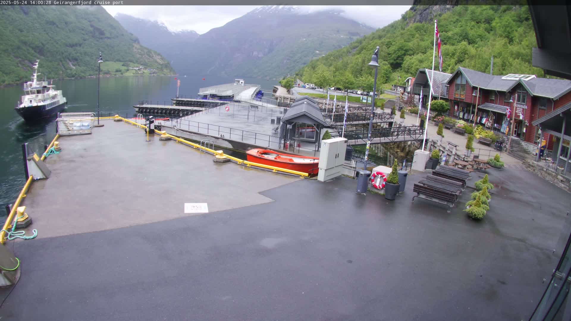 A cruise ship port in a fjord, with overcast weather, features a docked ferry, small boats, a paved area with benches, and buildings along a waterfront.