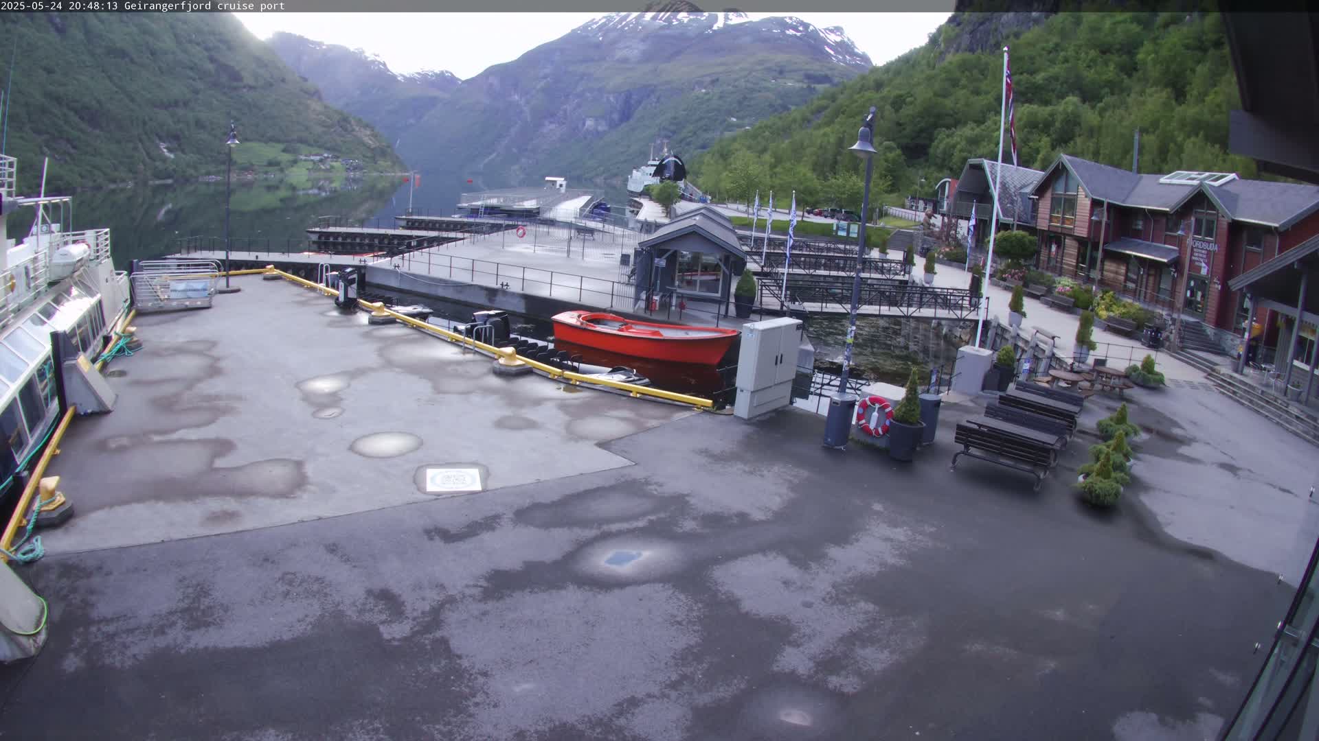 A wet cruise ship port with a red boat, several benches, and buildings nestled between mountains, under an overcast sky.