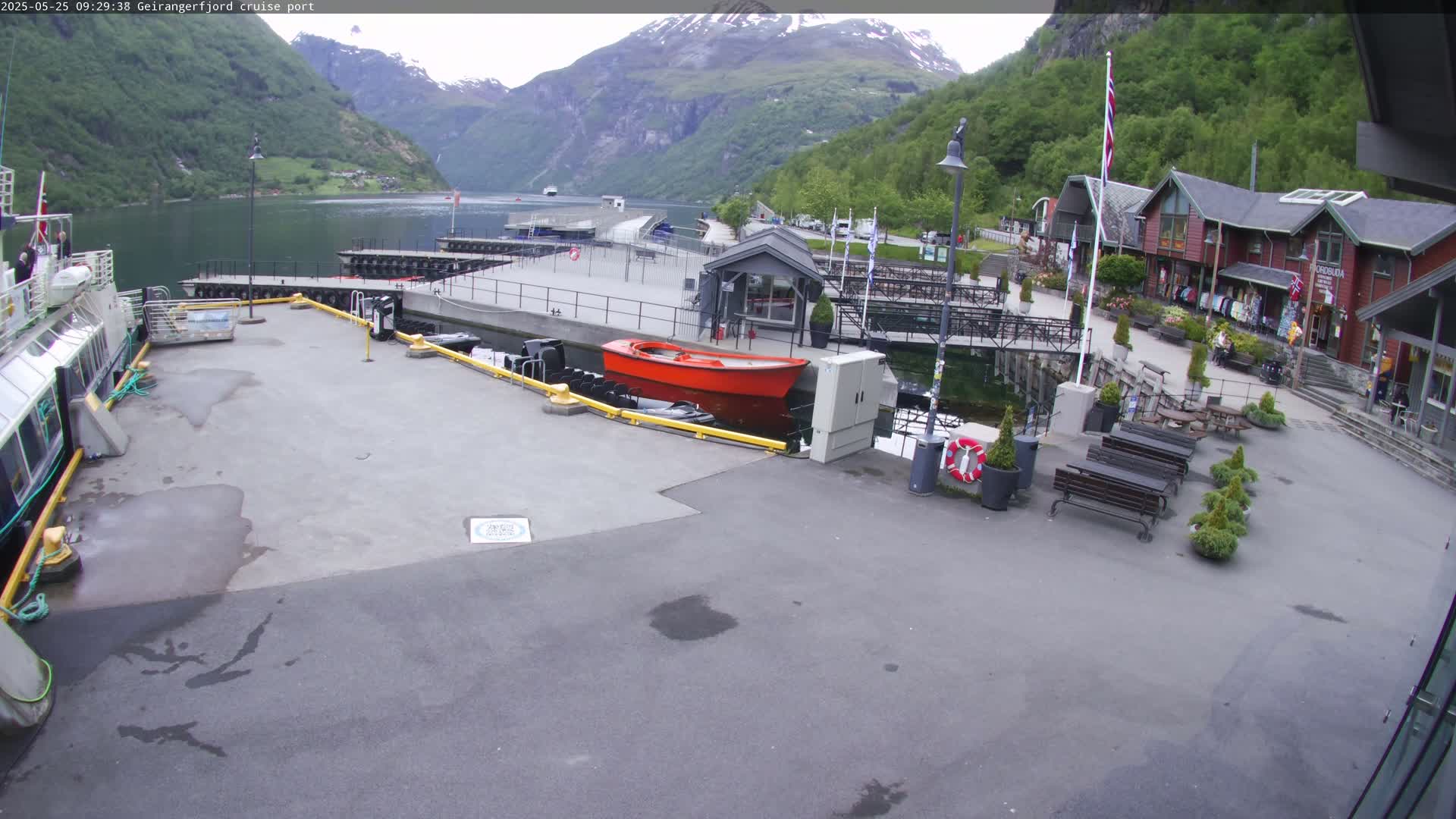 A cruise ship port in a fjord, under partly cloudy skies, with a red boat docked and mountains in the background.