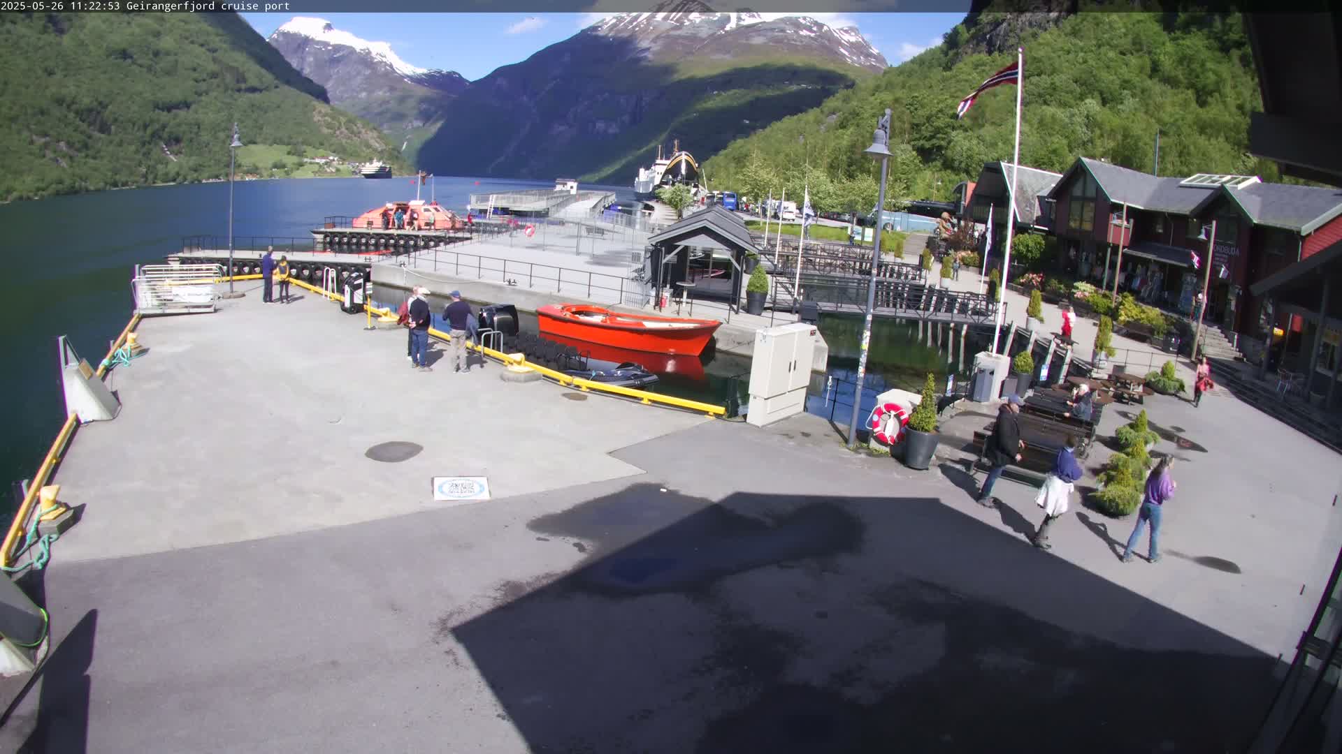 A cruise port on a sunny day, nestled between mountains, shows several people walking around the dock with a few boats moored nearby.