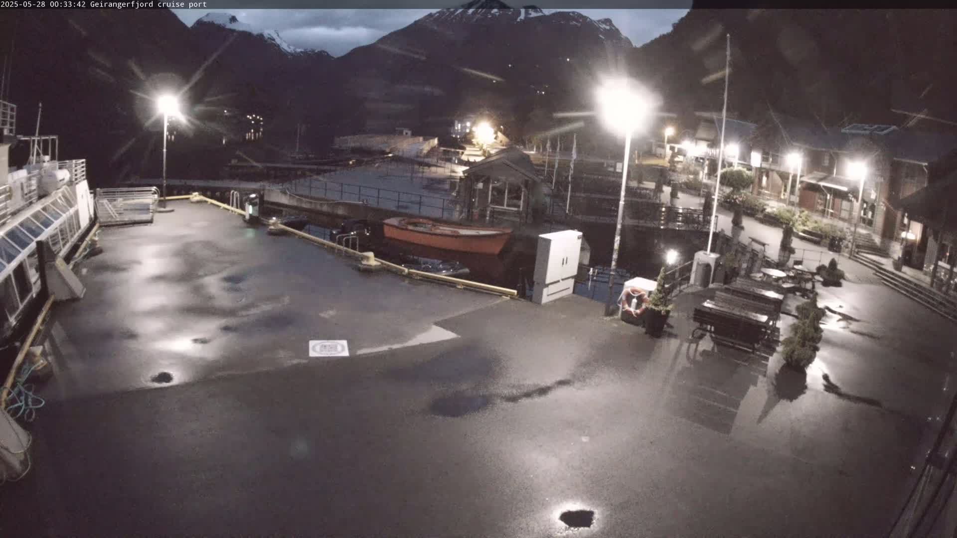A wet, nighttime scene at a cruise port shows a mostly empty dock with a cruise ship partially visible, several small boats, and buildings under overcast skies.