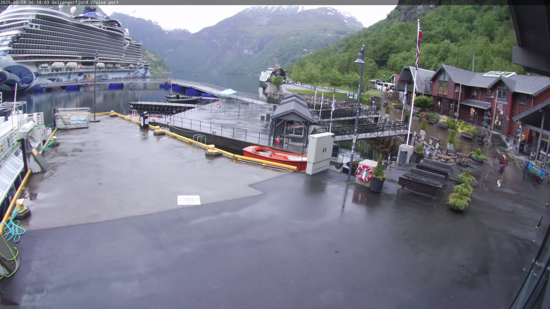 A cruise ship is docked at a port in a fjord surrounded by mountains under overcast skies, with rain-slicked surfaces on the dock and surrounding areas.