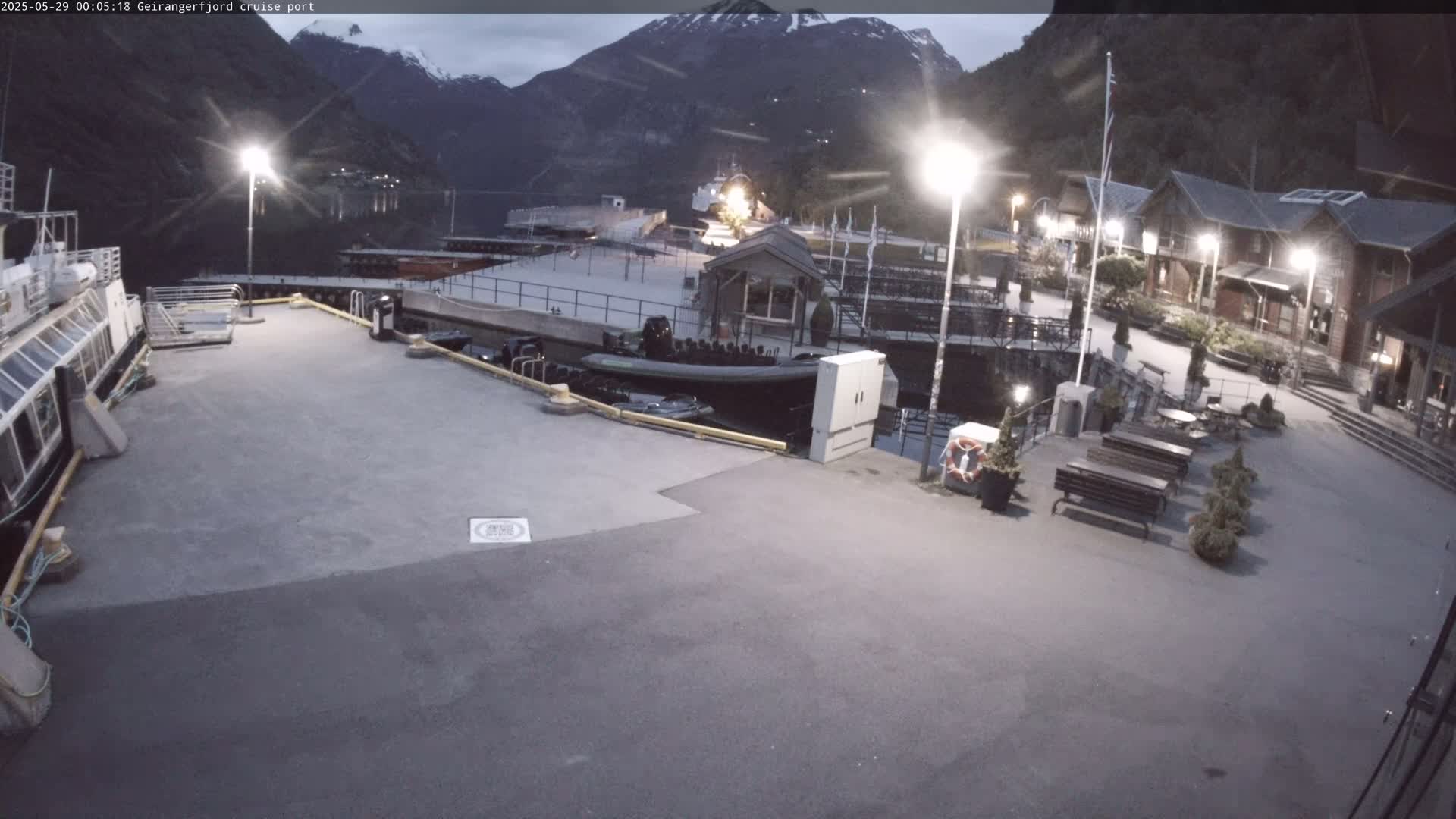 A cruise ship port at twilight, nestled between mountains, with several boats docked and benches in a paved area.