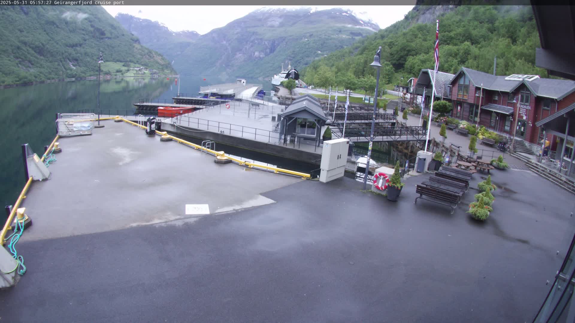 A cruise ship docks at a port nestled in a fjord, surrounded by green mountains under an overcast sky.