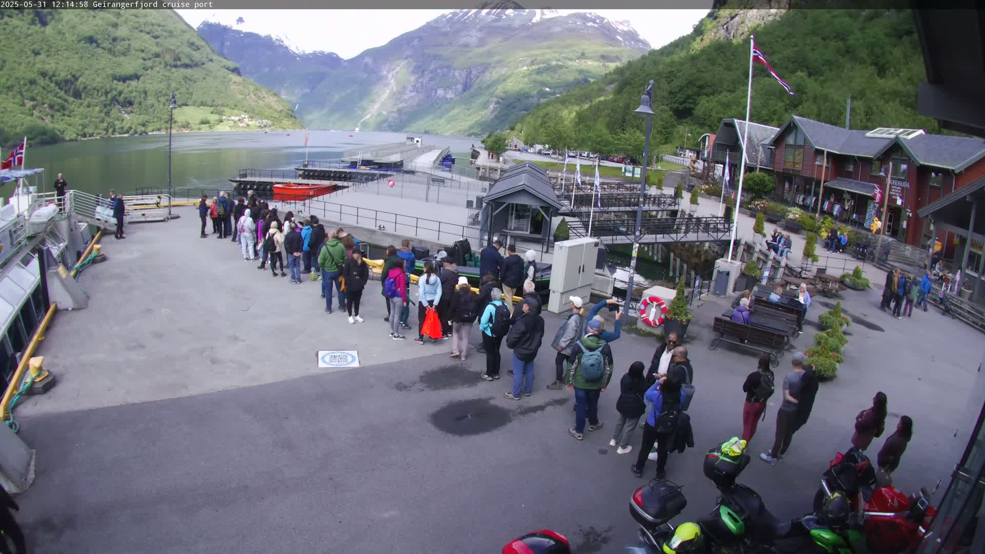 A crowd of people stands in lines at a cruise port in a fjord, under a partly cloudy sky, with mountains in the background.