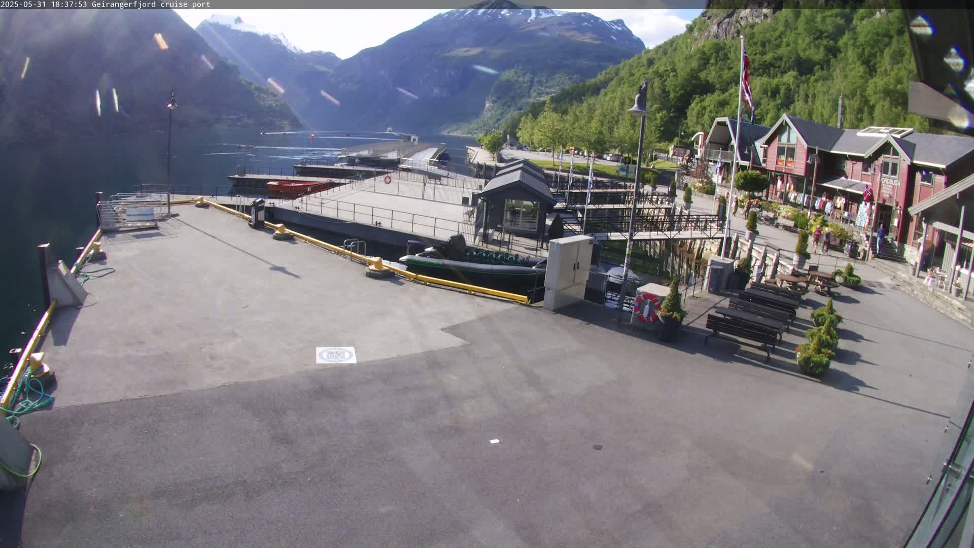 A sunny cruise ship port nestled between steep, green mountains overlooking a calm body of water, featuring docked boats, several buildings, and outdoor seating areas.
