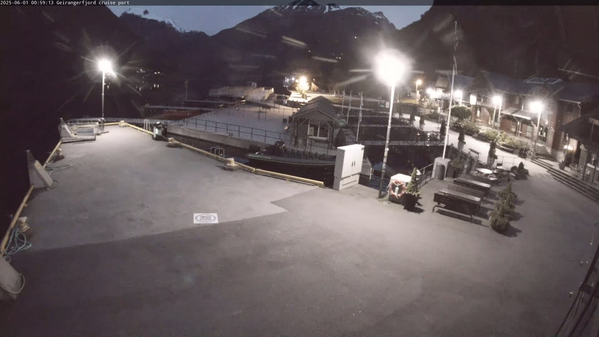 A nighttime view of an empty cruise port, illuminated by several bright lights, with a small boat docked near a building, set against a dark mountain backdrop.