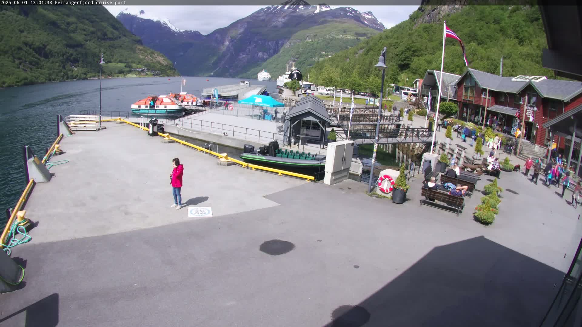 A cruise ship port in a fjord, under sunny skies, shows several boats docked, people relaxing on benches, and a person standing near the water's edge.