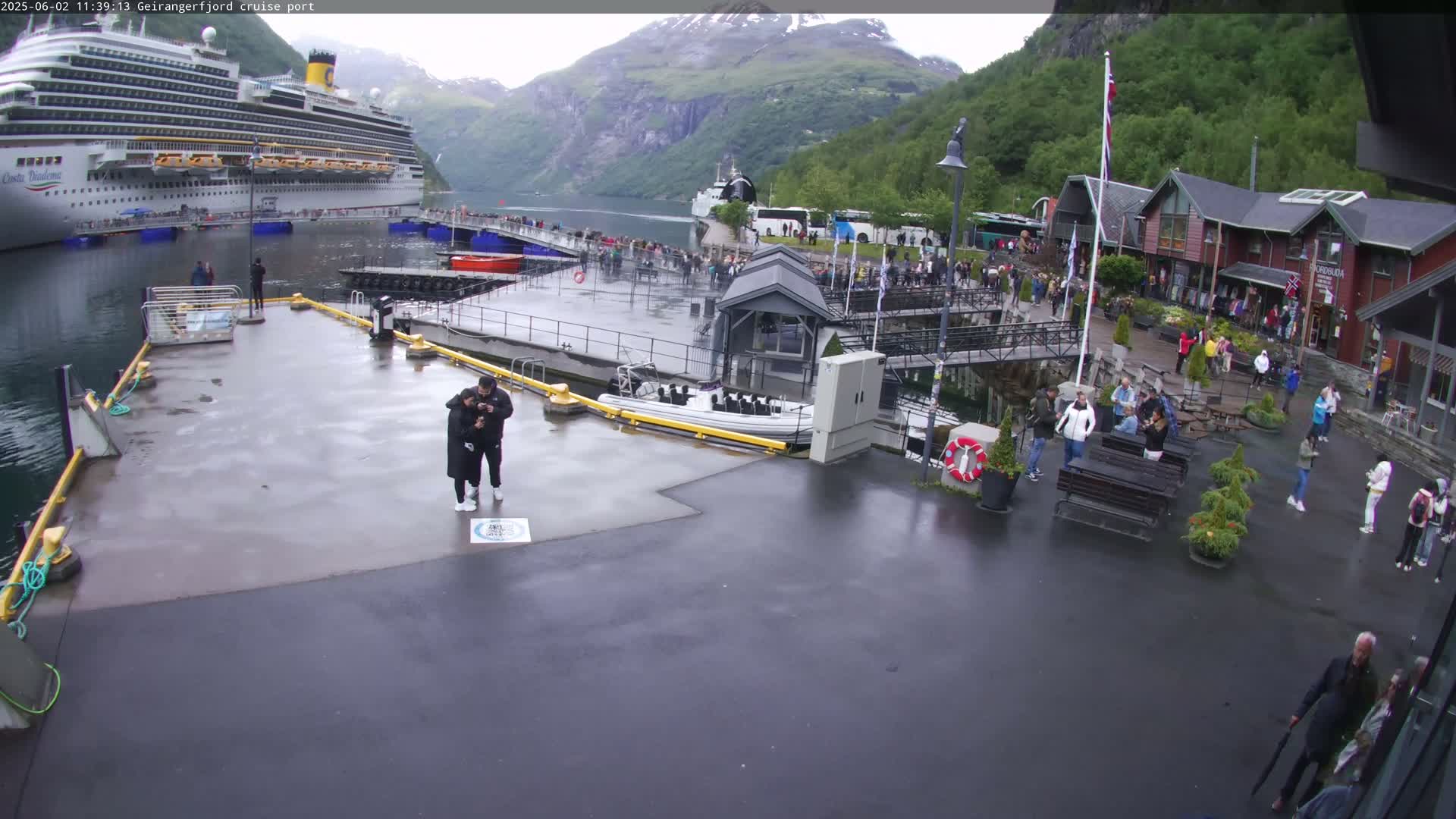 A cruise ship is docked at a port in a fjord, with overcast weather and many people gathered on the docks and surrounding area.
