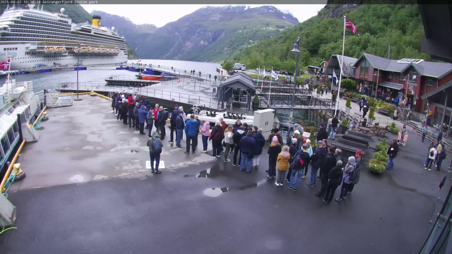 A large cruise ship is docked at a port in a fjord surrounded by mountains, with many people standing in groups on the dock under overcast skies.