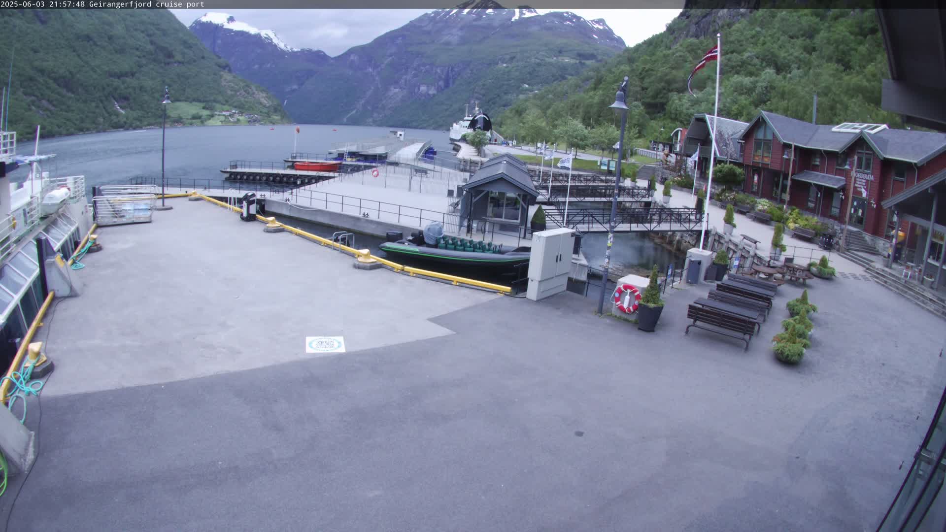 A cruise ship port in a fjord, under an overcast sky, features several small buildings, docked boats, and outdoor seating areas.