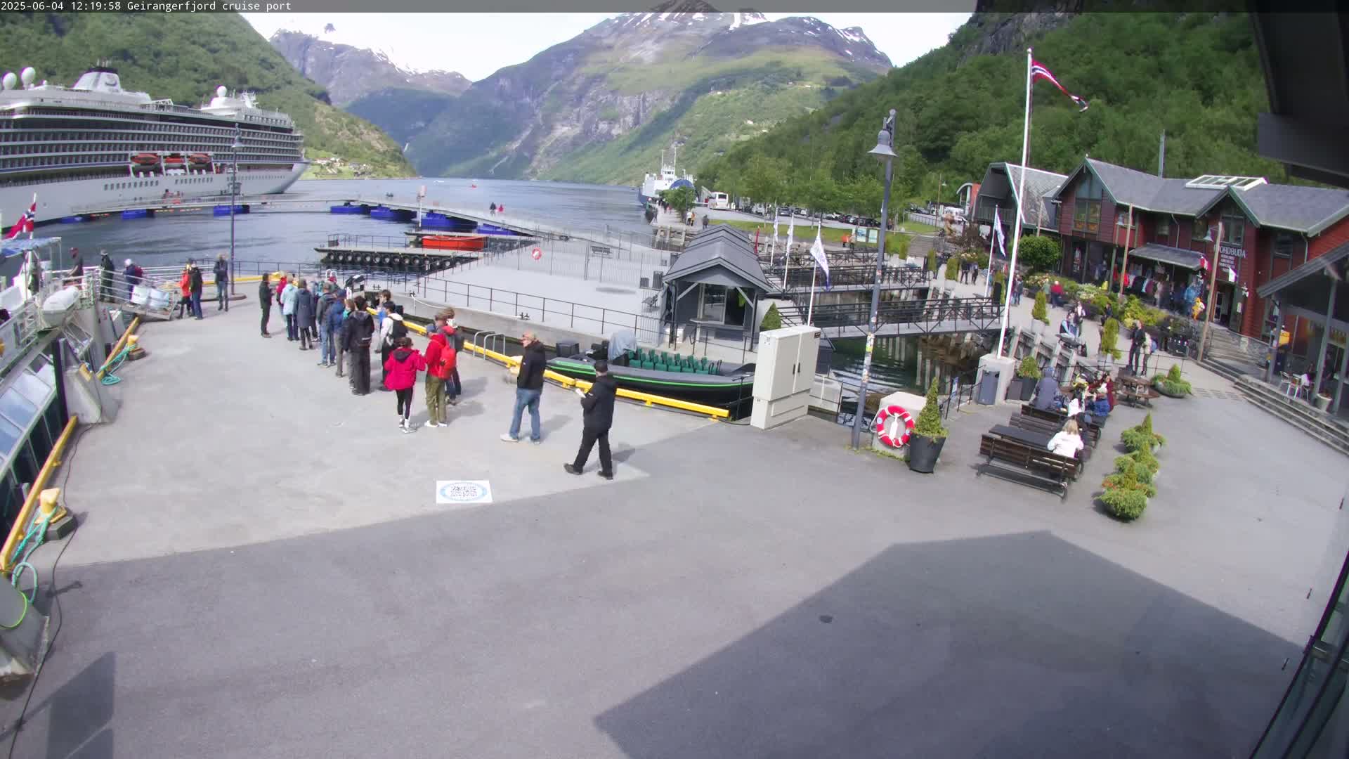 A cruise ship is docked at a port surrounded by mountains and a group of people are gathered on the dock on a sunny day.