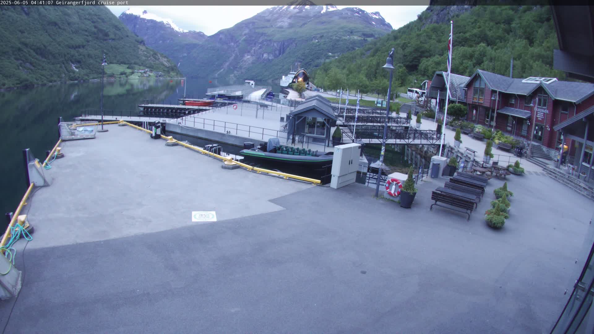 A cruise ship port in a fjord, under an overcast sky, features several docked boats, buildings, and outdoor seating areas.