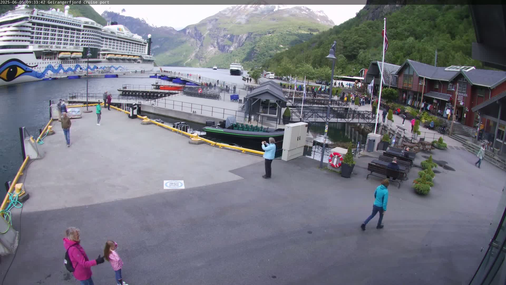 A cruise ship is docked at a port in a fjord surrounded by mountains under a partly cloudy sky, with several people walking around the area and a smaller boat moored nearby.