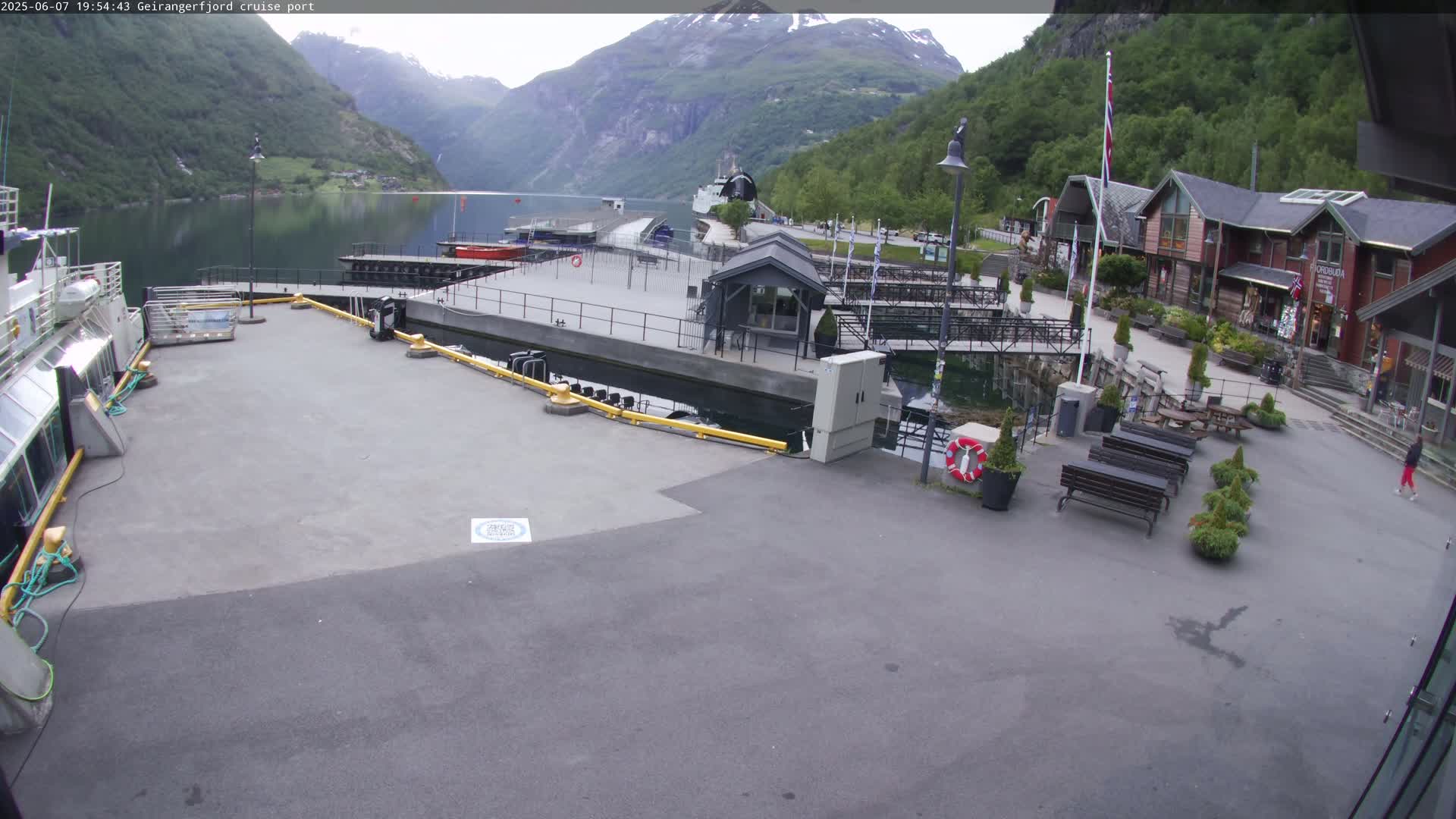 A cruise ship port in a fjord, with several buildings, benches, and a calm body of water surrounded by mountains under an overcast sky.