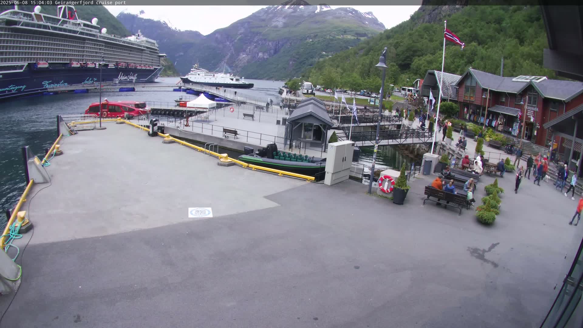 A cruise ship is docked at a port next to smaller vessels and buildings, with people walking around on a mostly sunny day in a fjord setting.