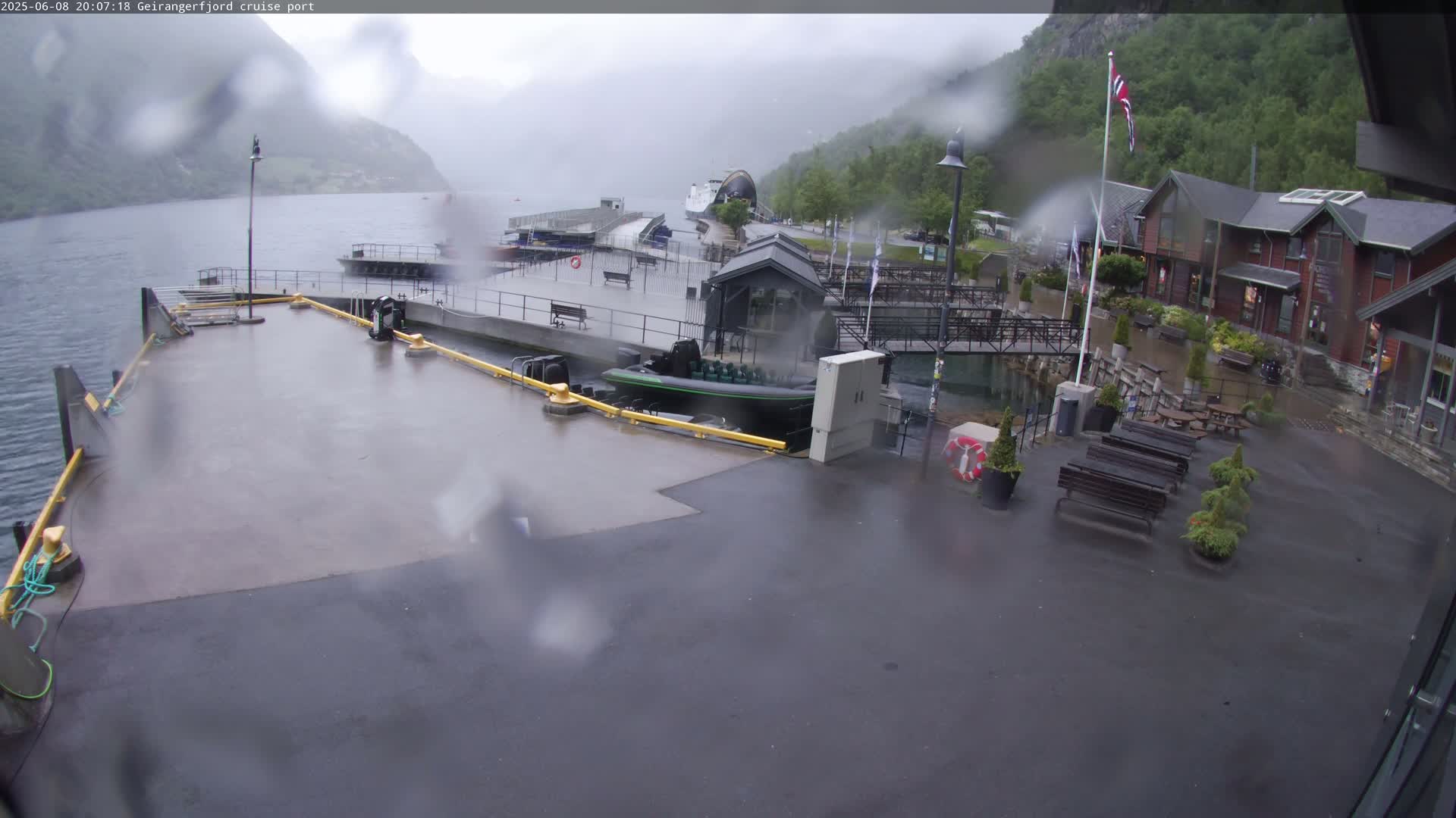 A rainy view of a cruise port, showing a dock with a small boat, several buildings, and a calm body of water surrounded by mountains in the background.